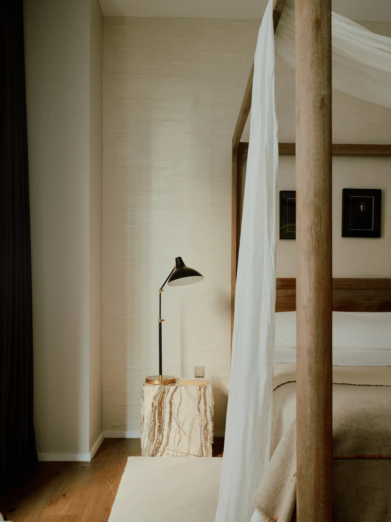 A minimalist bedroom featuring a wooden four-poster bed with a marble base and draped white curtains, alongside a side table