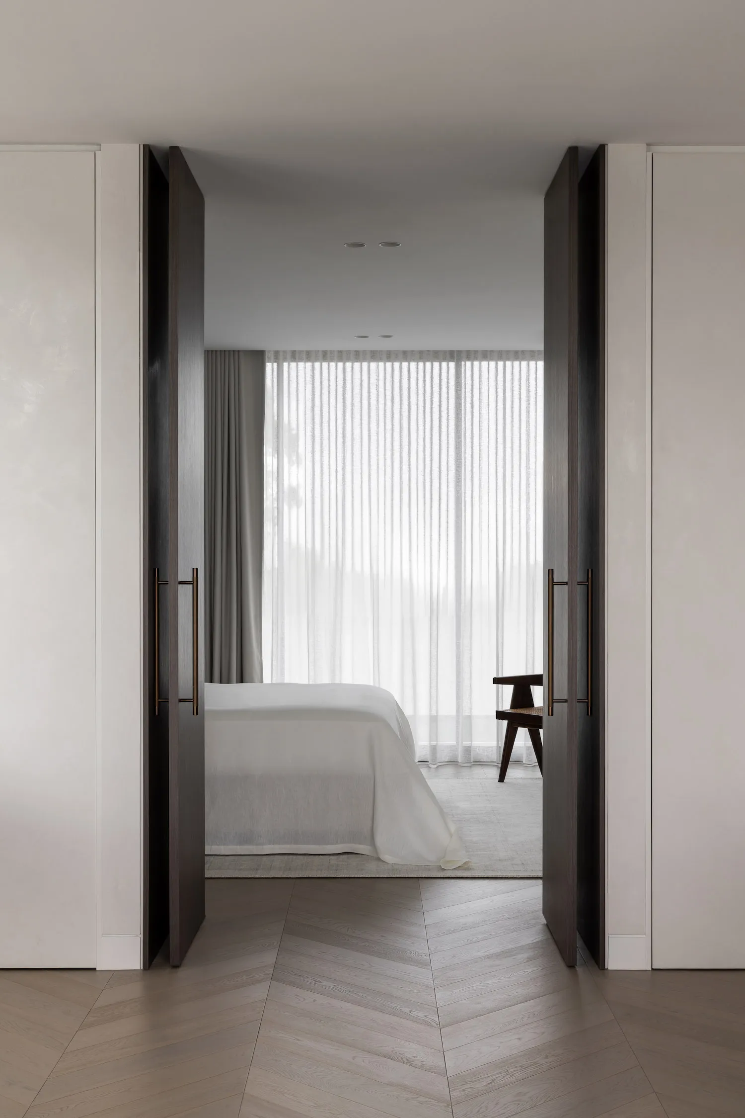 A minimalist bedroom with a bed dressed in white sheets, a patterned floor, and large windows dressed with white curtains.
