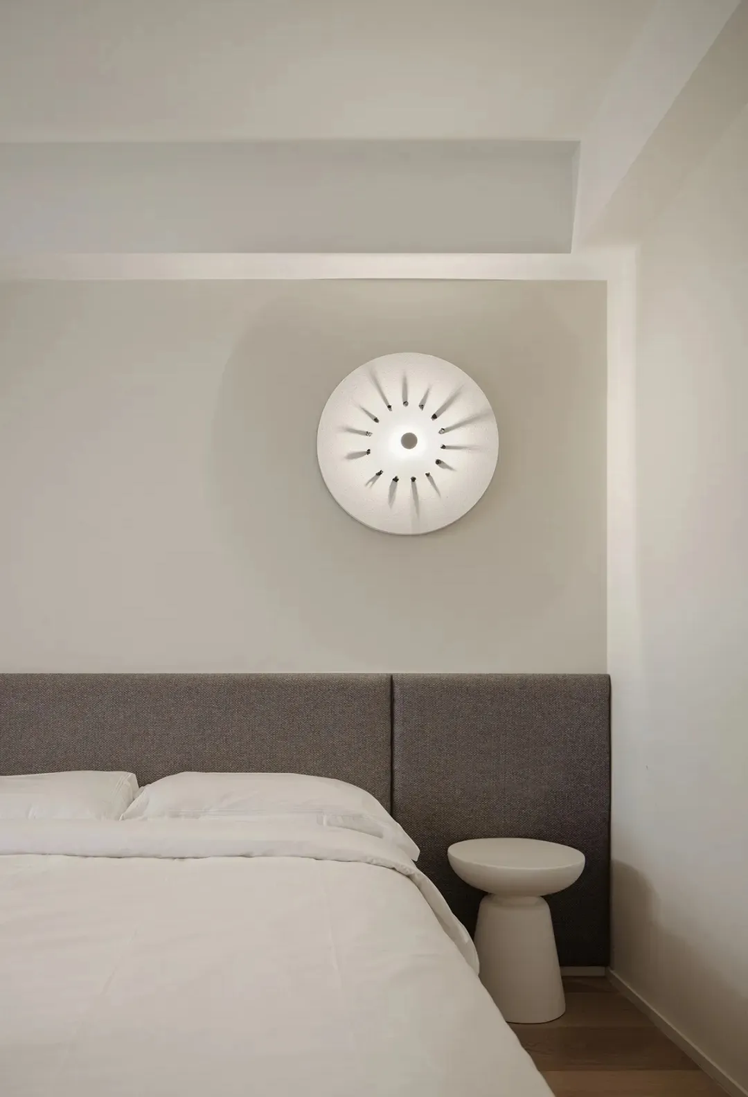 A minimalist bedroom featuring a white bed with a gray upholstered headboard against a light-colored wall, illuminated by a u