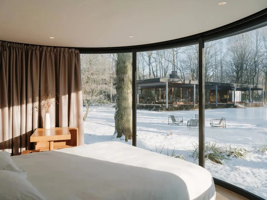 A minimalist bedroom with a white bed, glass windows overlooking a snowy landscape, and modern furnishings.