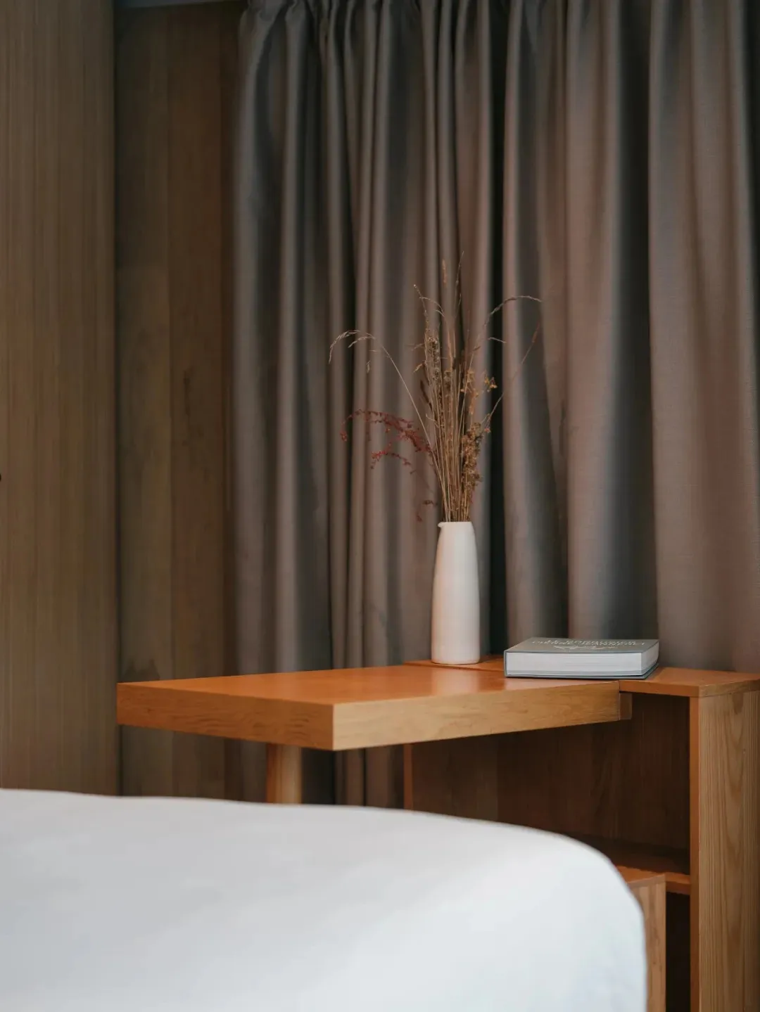 A minimalist bedroom featuring a wooden side table with a vase of dried flowers and a book.