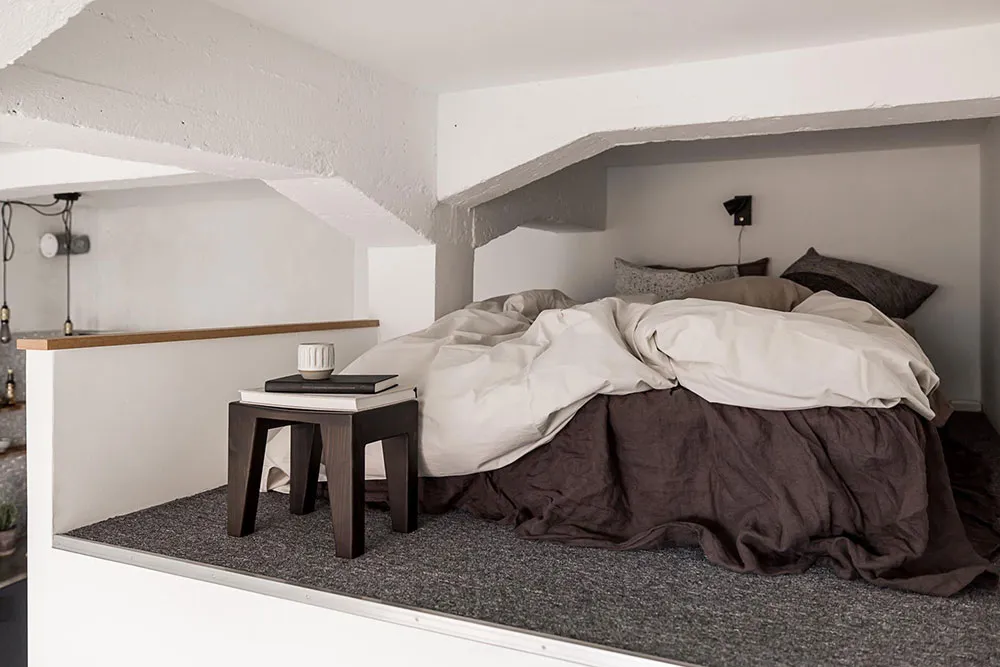 A minimalist bedroom with a bed covered in beige and brown bedding, a wooden bedside table, and concrete walls.