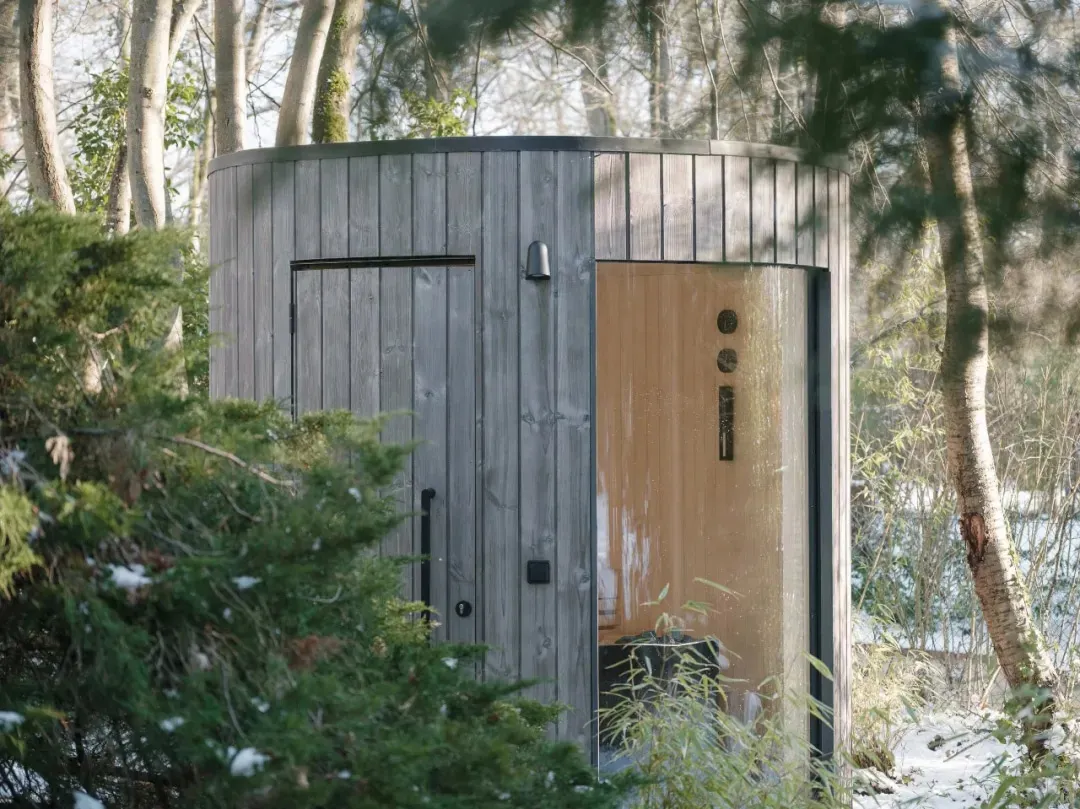 A modern outdoor spa with a wooden exterior surrounded by trees and snow-covered ground.