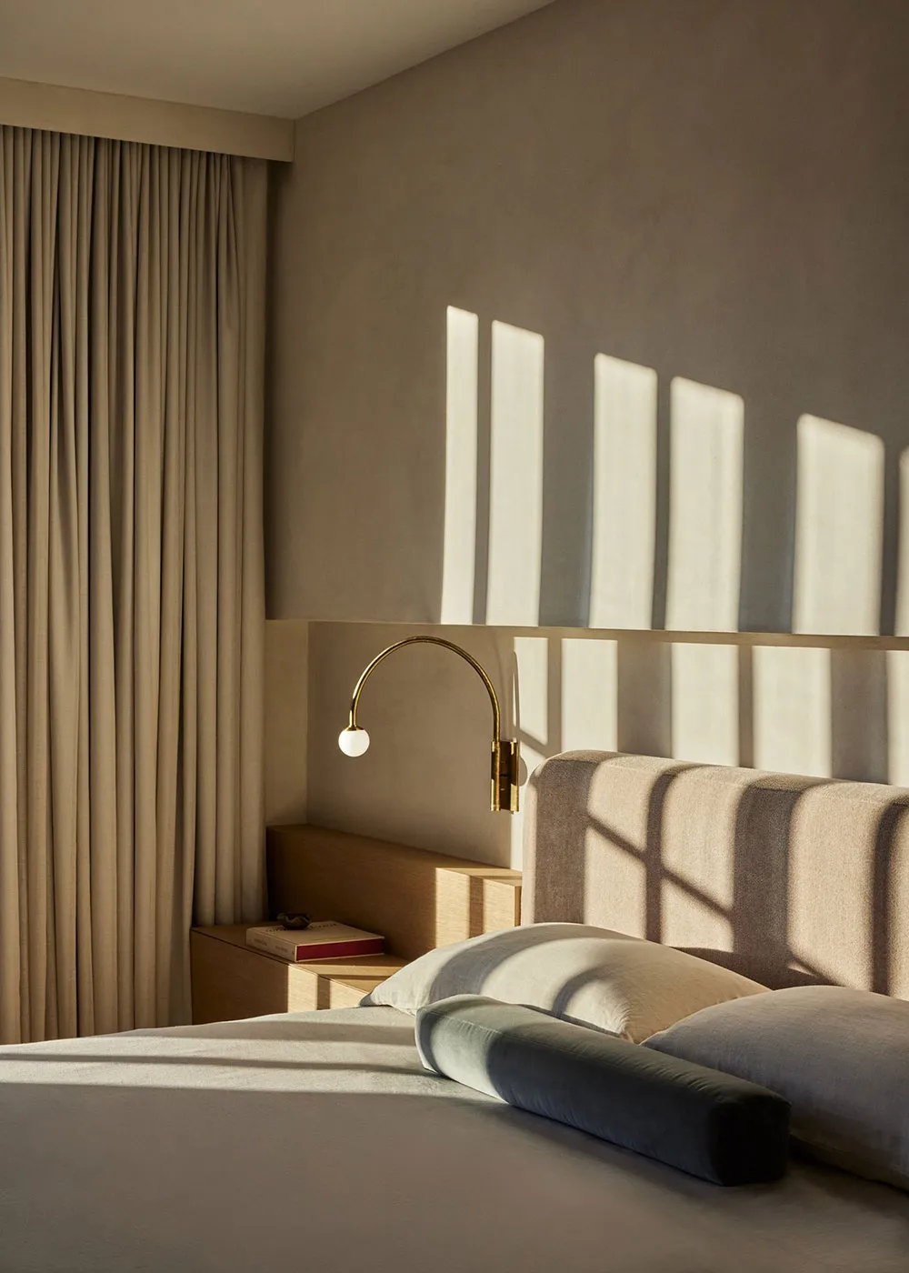 A minimalist bedroom with a bed, pillows, and a wall-mounted light fixture casting shadows on the wall.