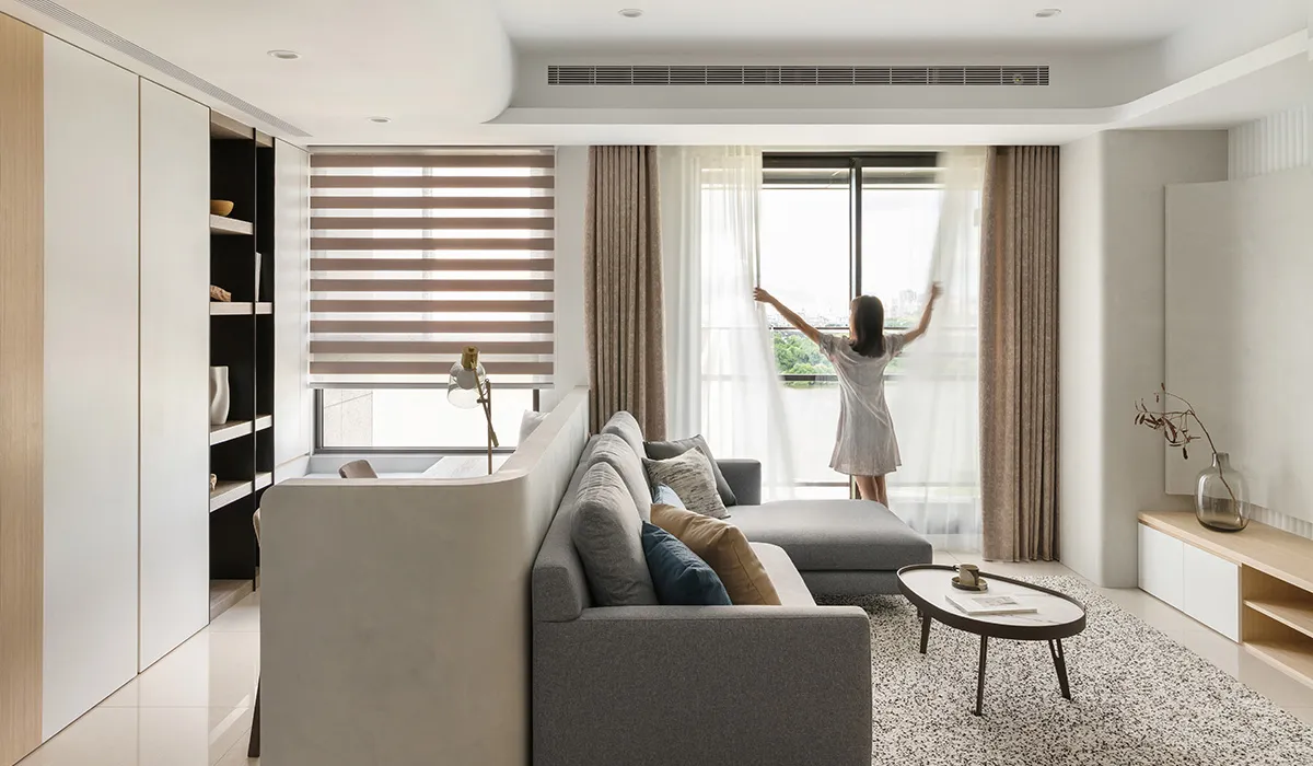 Person adjusting beige curtains in a minimalist living room with a gray sectional sofa and a round coffee table.