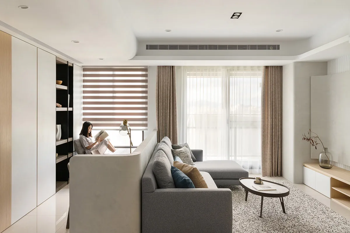 A minimalist living room with fabric curtains, concrete cabinets, and grey sofa, featuring a woman reading a book.