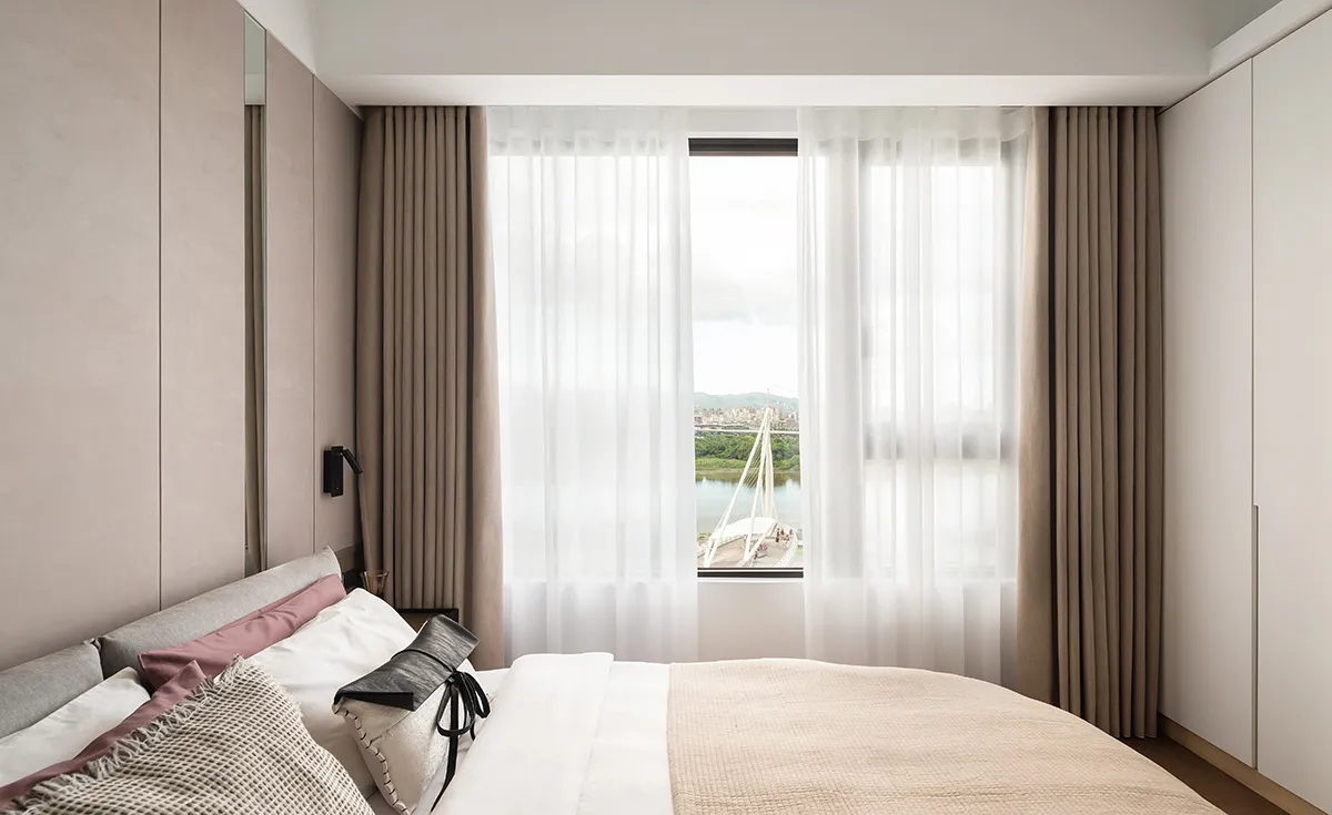 A serene bedroom featuring modern design with light-colored fabric and a glass window overlooking a scenic view.