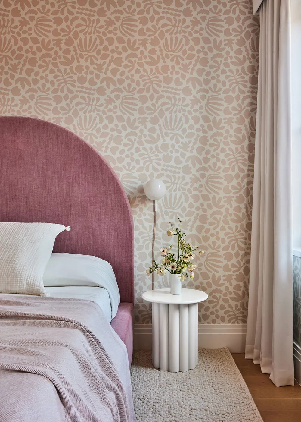 A modern bedroom featuring a bed with a pink upholstered headboard, a white side table with a vase of flowers, and patterned 