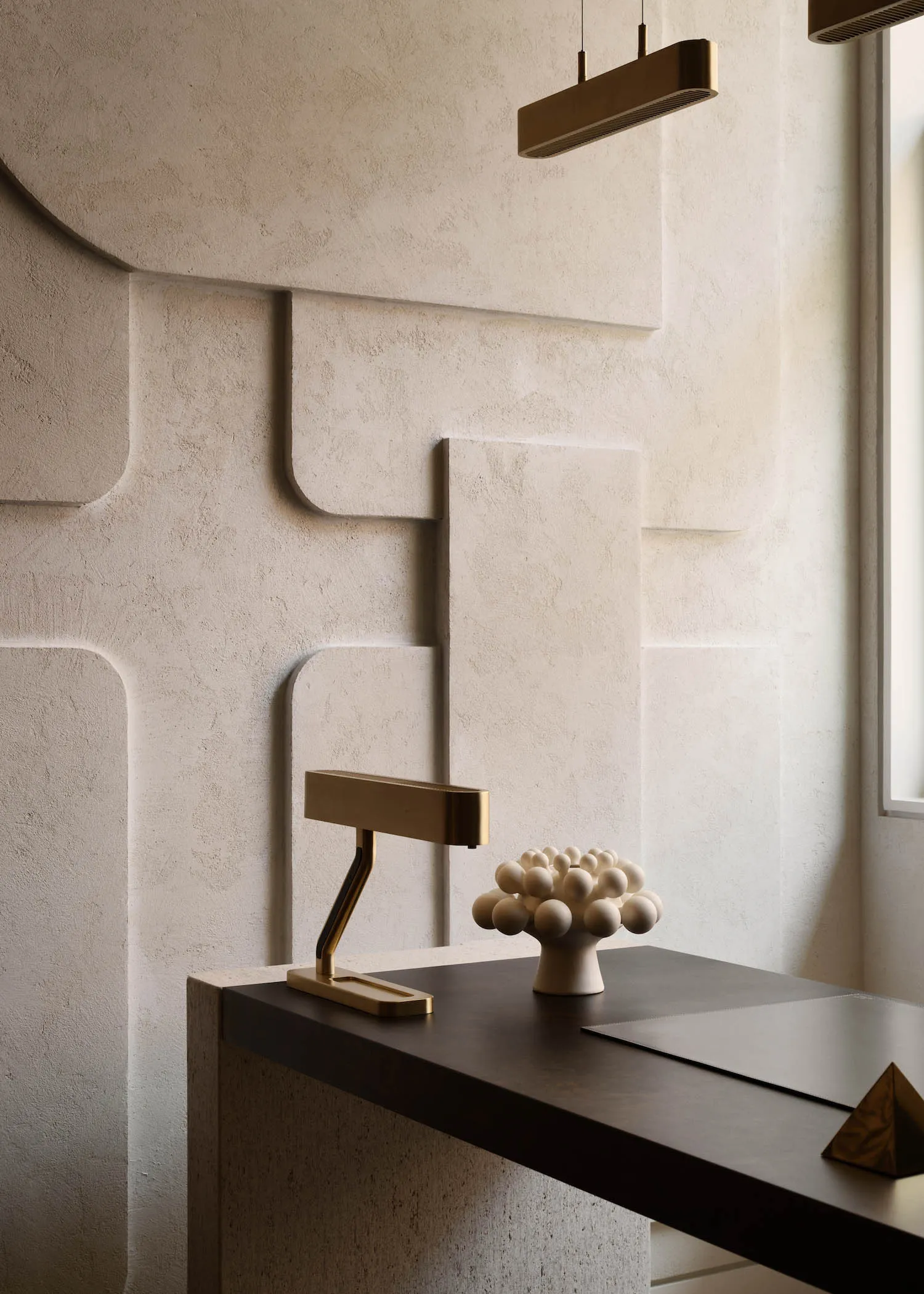 A minimalist workspace with a concrete and wood desk, a brass desk lamp, and a decorative sculpture on a textured wall.