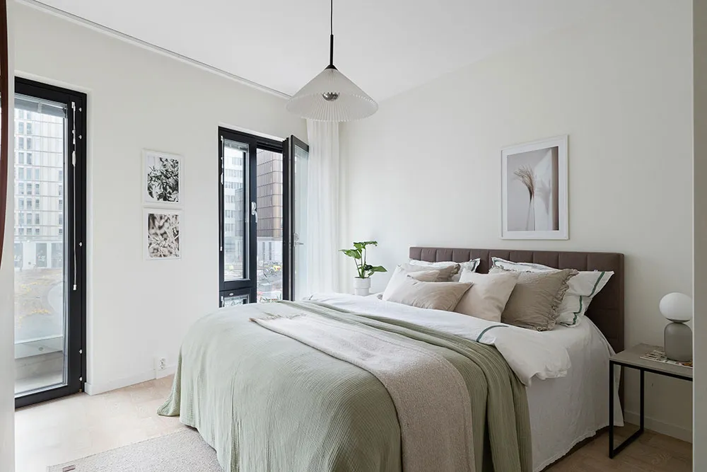 A modern bedroom with light-colored furniture and green bedding, featuring a large window with city views and a minimalist de