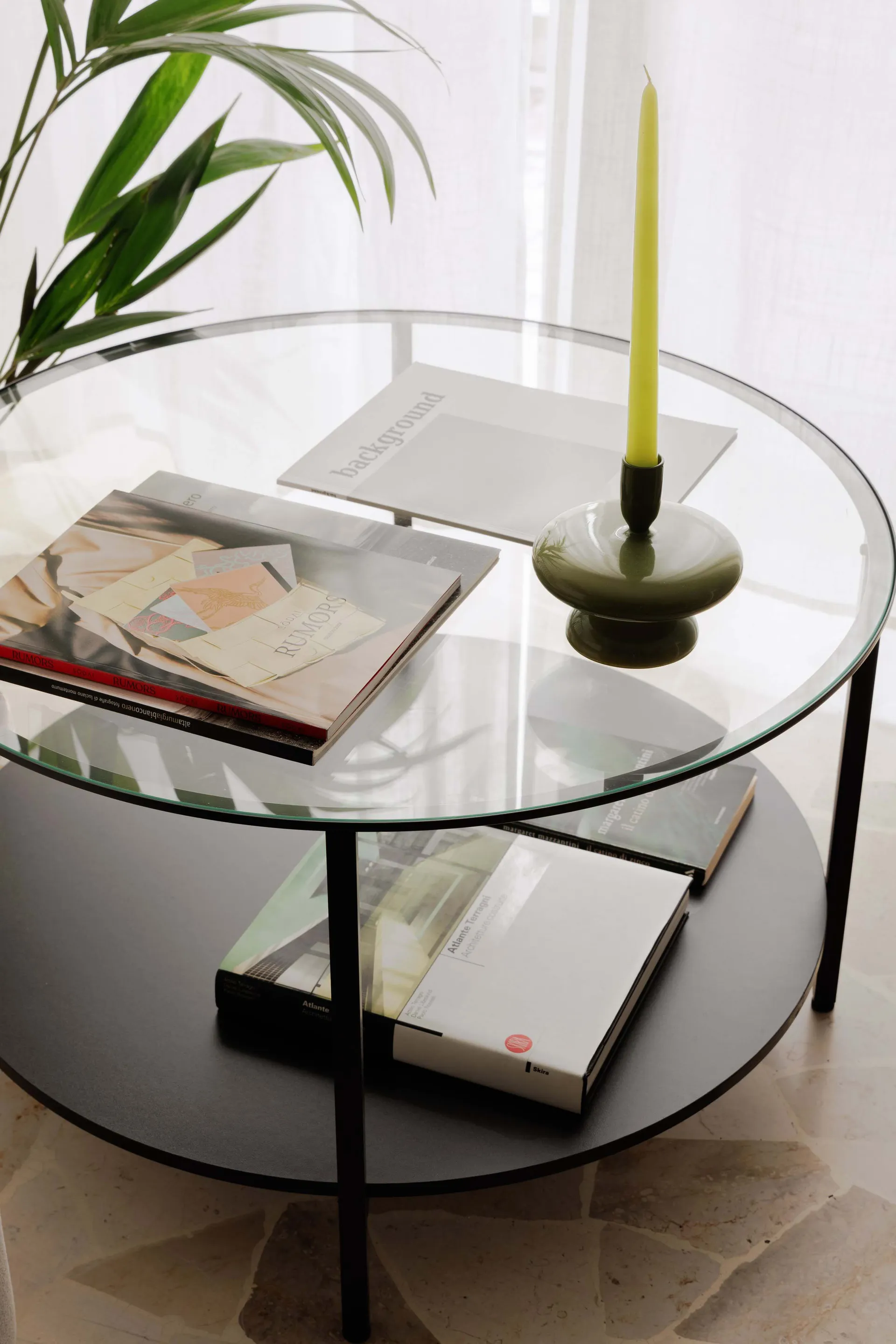 A modern coffee table made of glass and metal with books and a candle.