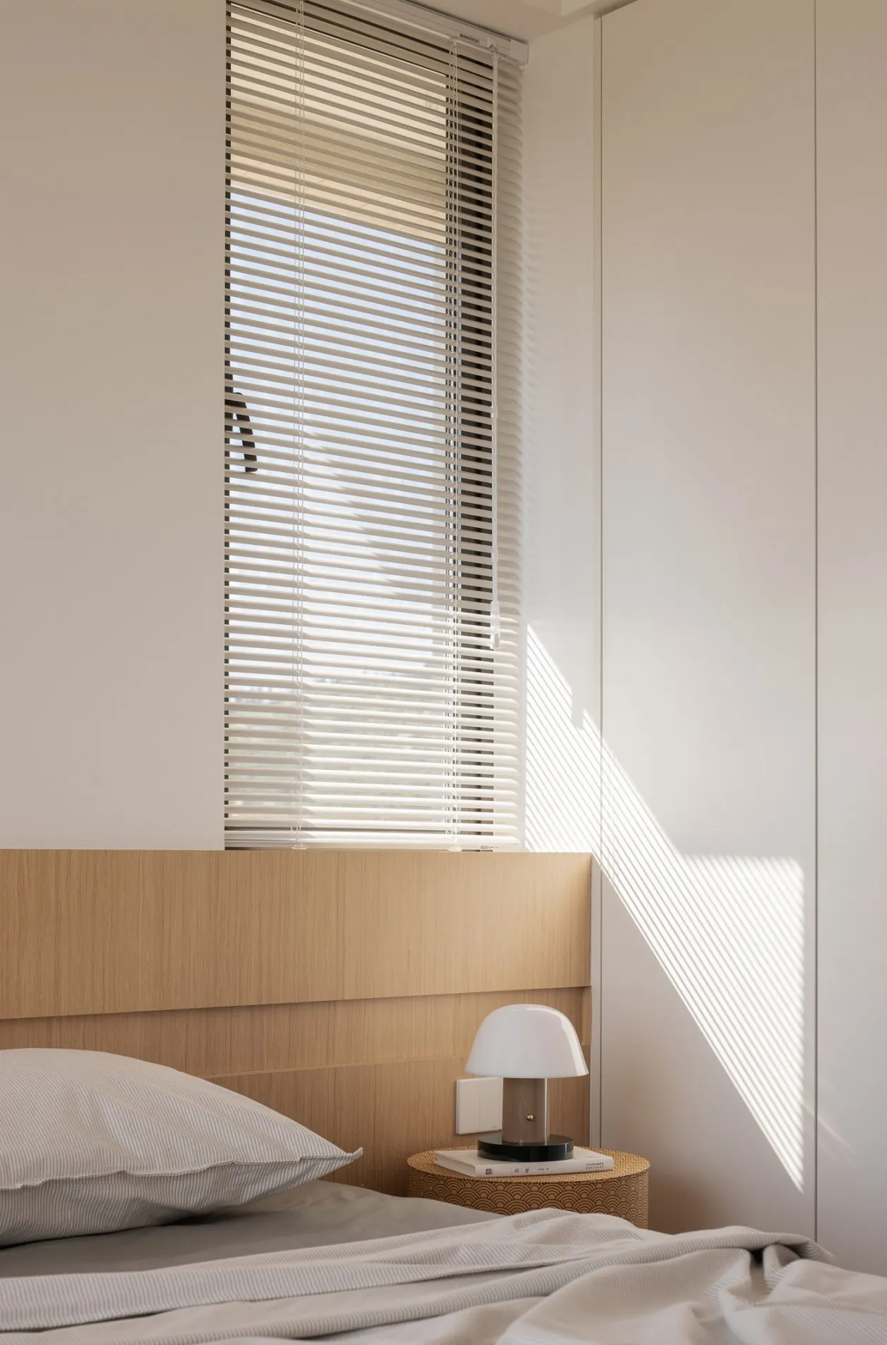 A minimalist bedroom featuring a bed with wood headboard and blinds, illuminated by sunlight casting geometric shadows.