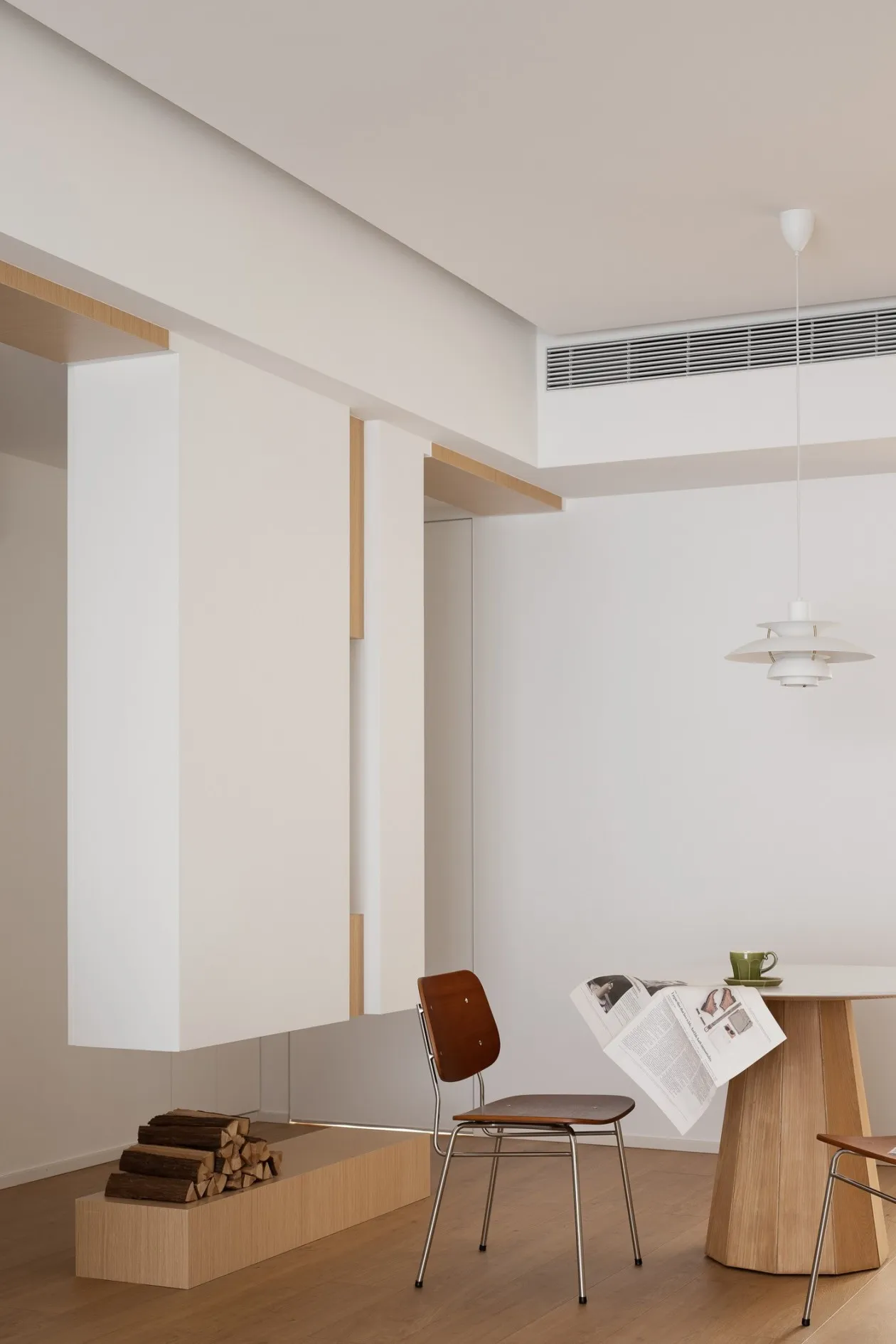 A minimalist living room with white walls, wooden furniture, and a metal pendant light hanging above a wooden dining table.