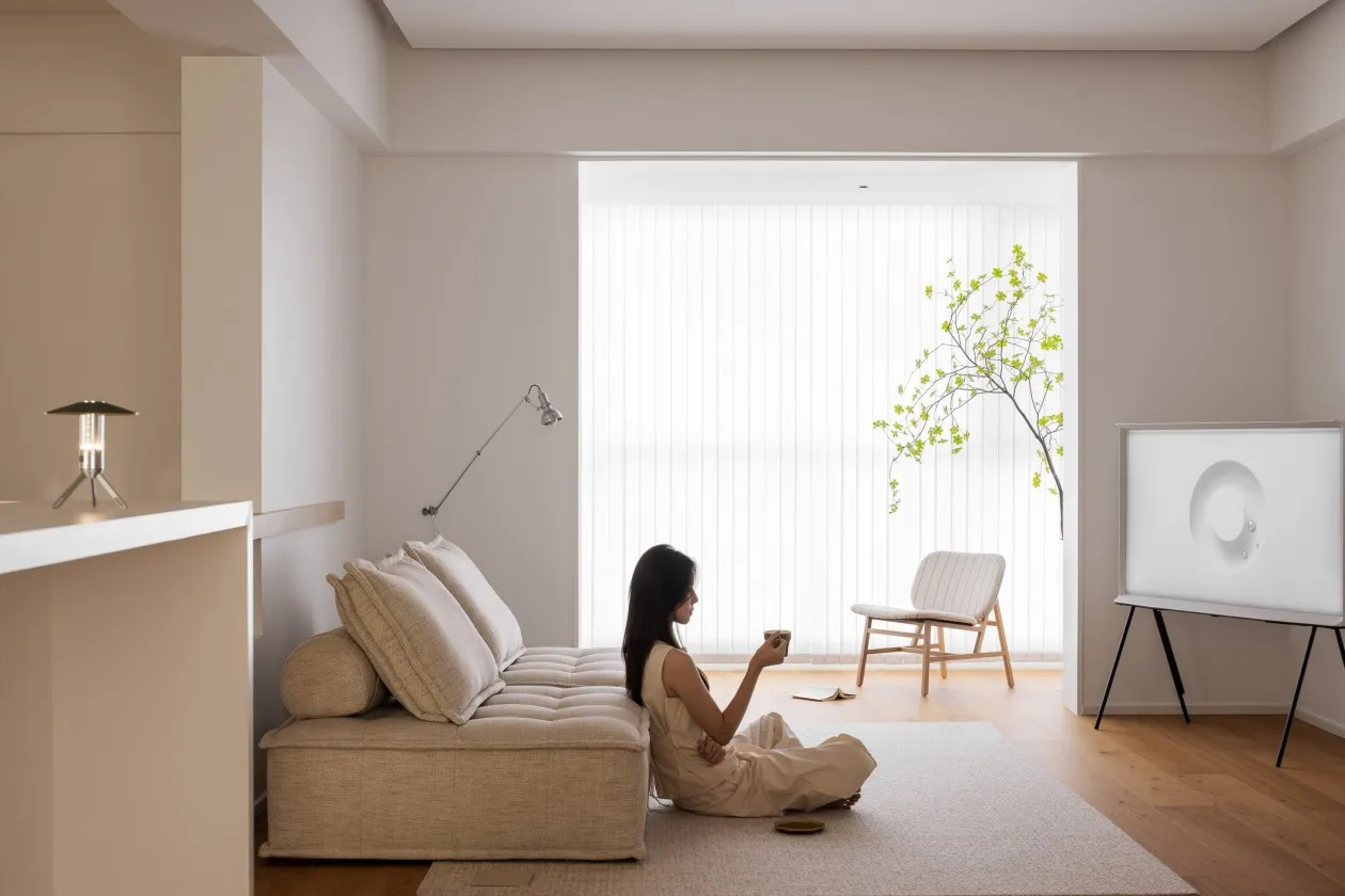 A minimalist living room with a beige fabric sofa, a tree by the window, and a woman sitting on the floor with a drink.