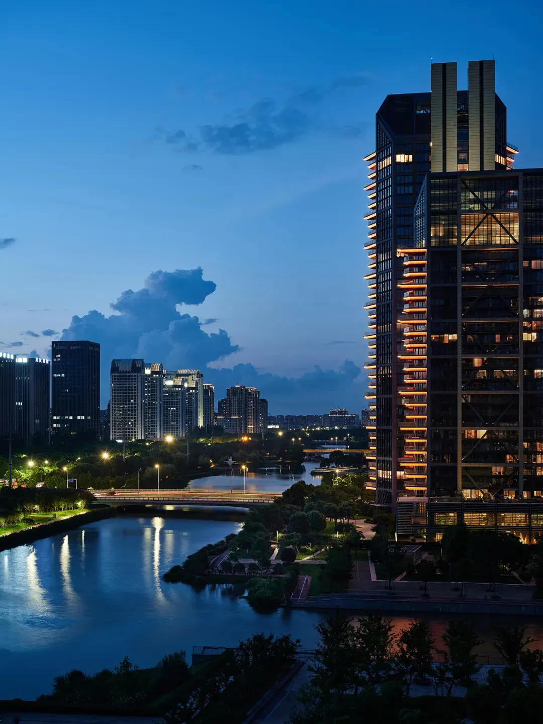 A modern cityscape featuring illuminated skyscrapers and a calm river at dusk.