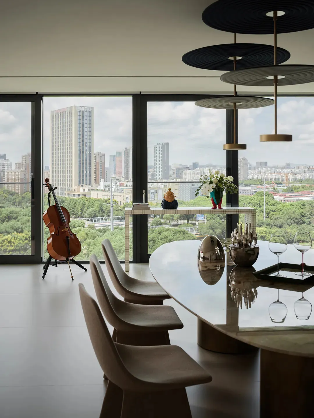 Modern dining room featuring a glass table, wooden chairs, and metal pendant lights, overlooking a cityscape through a large 