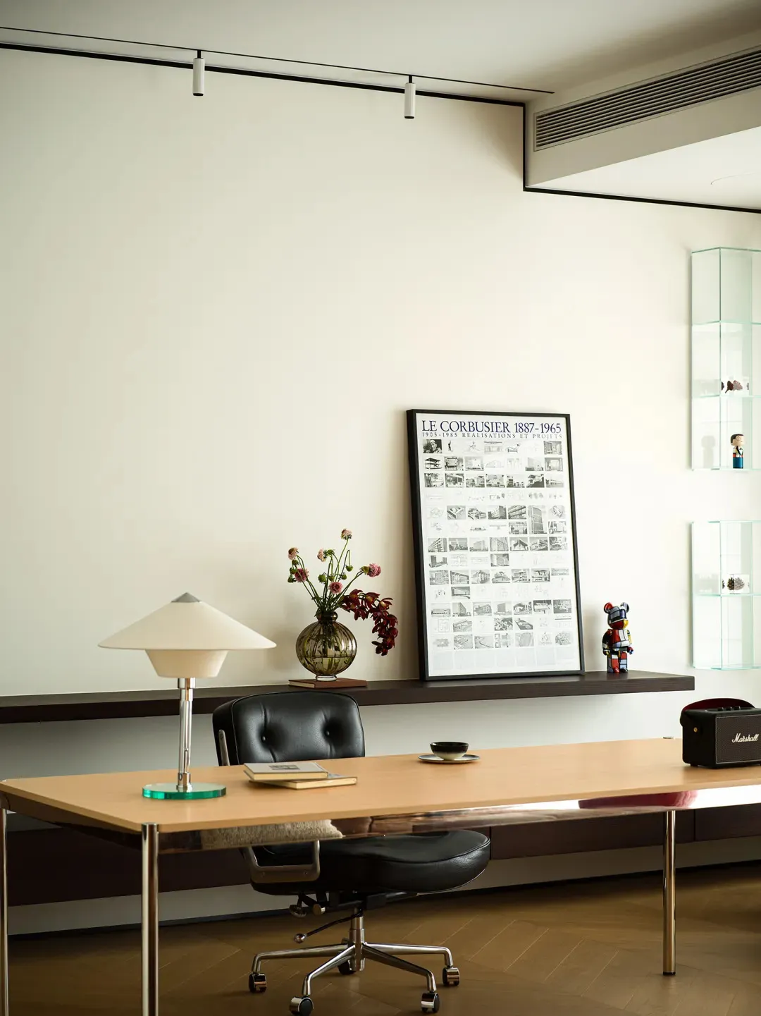 A minimalist office desk with a wooden surface, a black swivel chair, a lamp, a vase with flowers, and a framed architectural