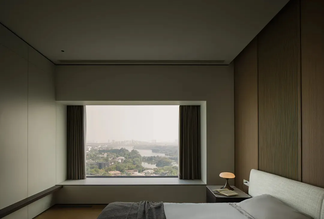A modern bedroom with brown and neutral colors, featuring a large window showcasing a cityscape with a river and trees.