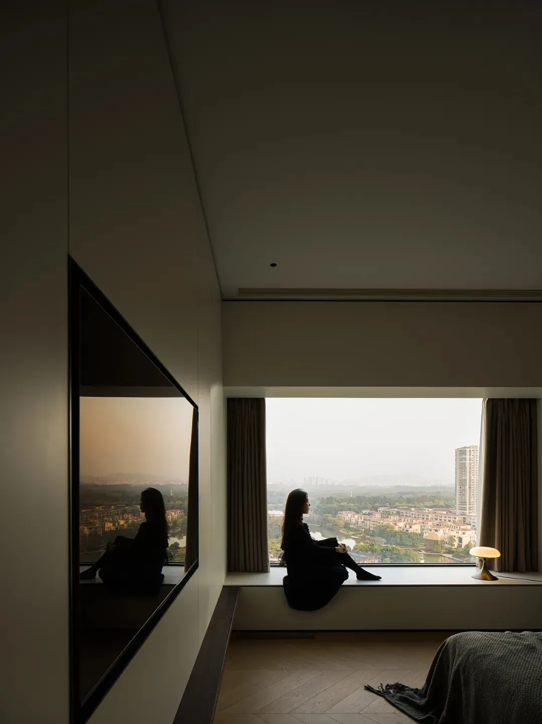 A person sitting on a window sill in a modern interior with glass walls, wooden flooring, and textile furnishings.