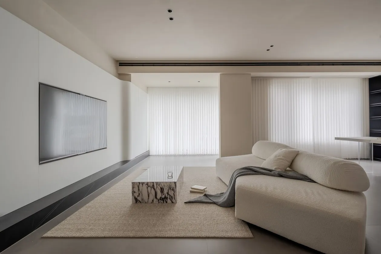 A minimalist living room with a neutral color palette, featuring a large white sofa, a marble coffee table, and a flat-screen