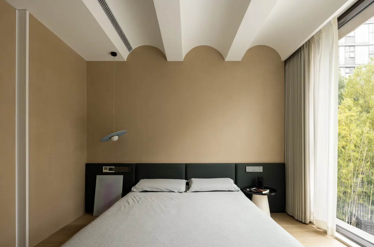A minimalist bedroom with a white sheet on the bed, beige walls, and a hanging light fixture.