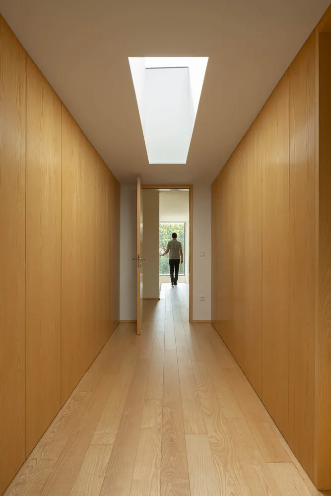 A minimalist wooden hallway with a skylight above a man walking towards a window.