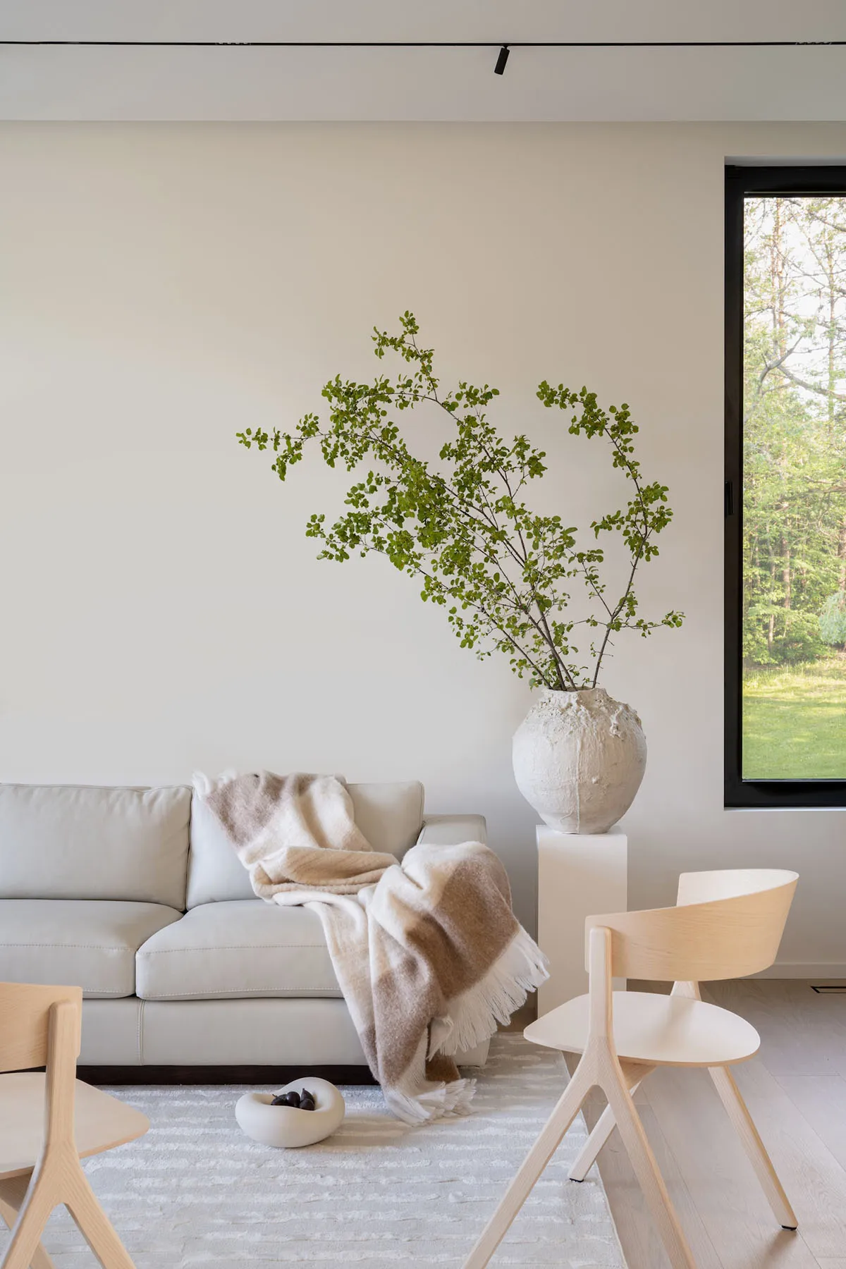 A minimalist living room with a beige sofa, a white vase with green branches, and light-colored wood chairs.