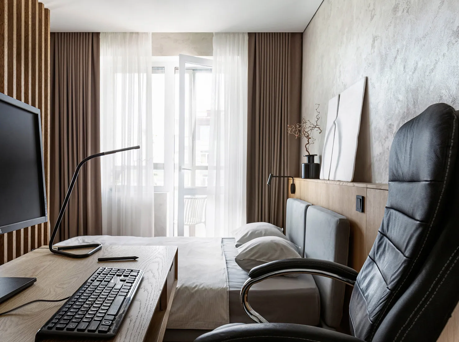 Modern minimalist bedroom with a black leather office chair, white bedspread, and white curtains by the large window.