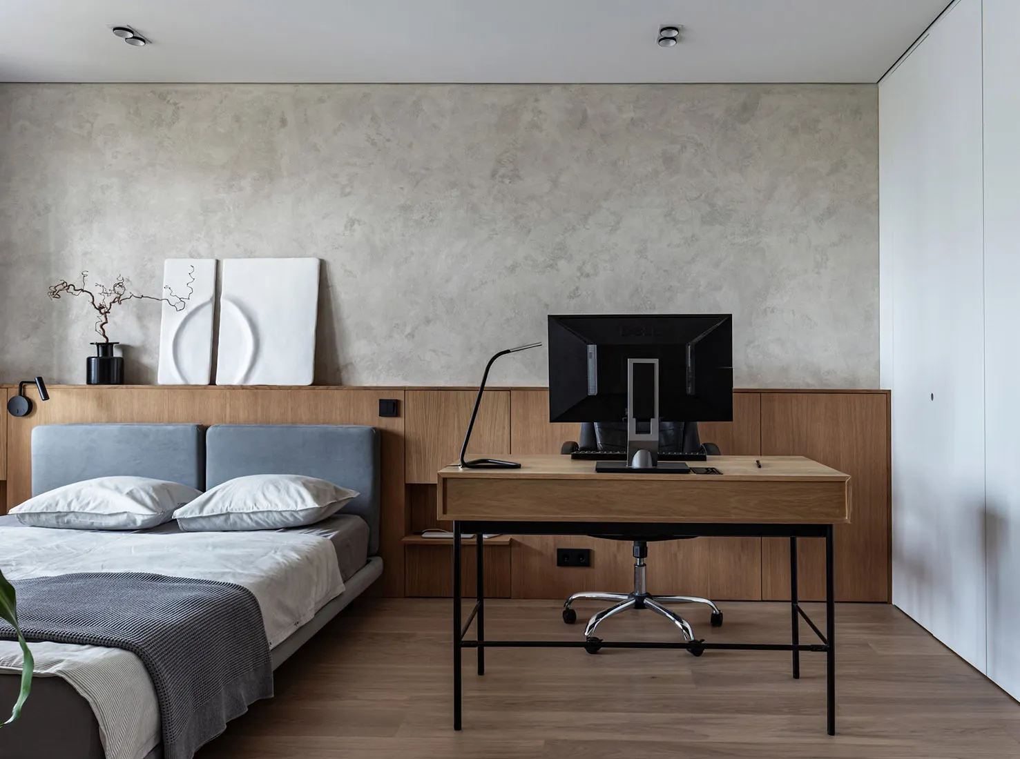 A modern minimalist bedroom featuring a wooden desk with a computer monitor, a bed with white and gray bedding, and a concret