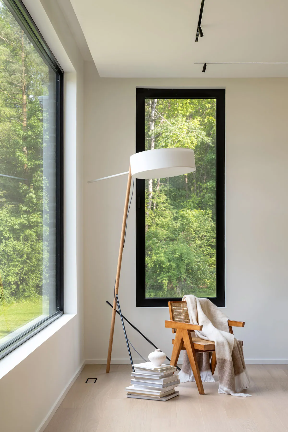 A minimalist living room with a modern floor lamp, books, and a wooden chair with a blanket next to large windows overlooking