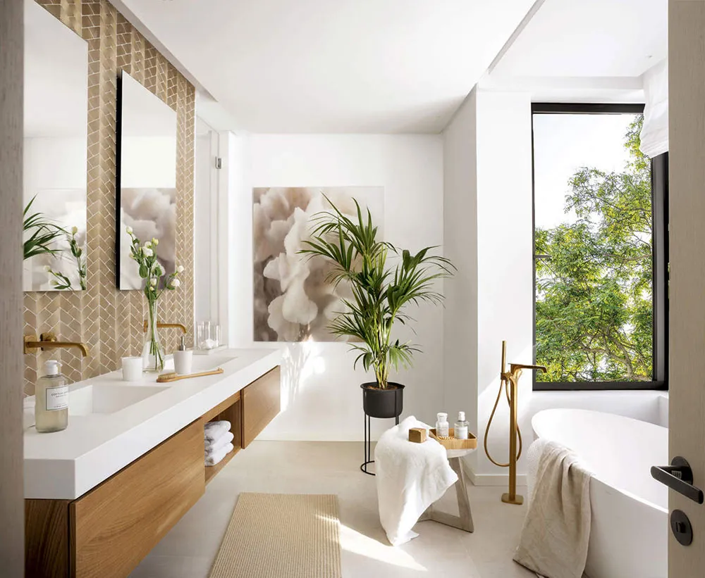 A modern bathroom with a white sink counter, wooden cabinets, a freestanding bathtub, and a large window showcasing greenery.
