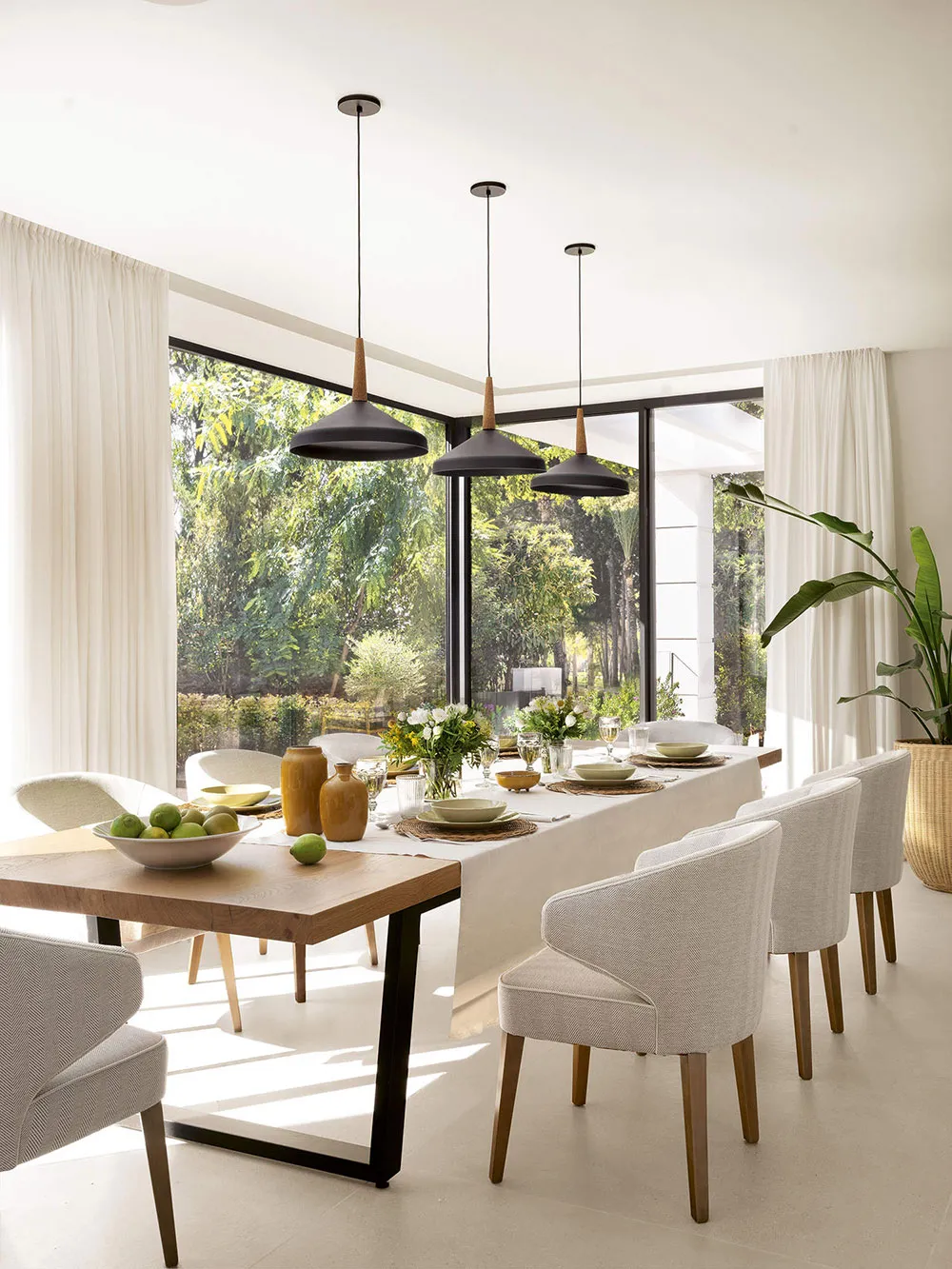 A modern minimalist dining room with large windows overlooking greenery, featuring a long dining table surrounded by white fa