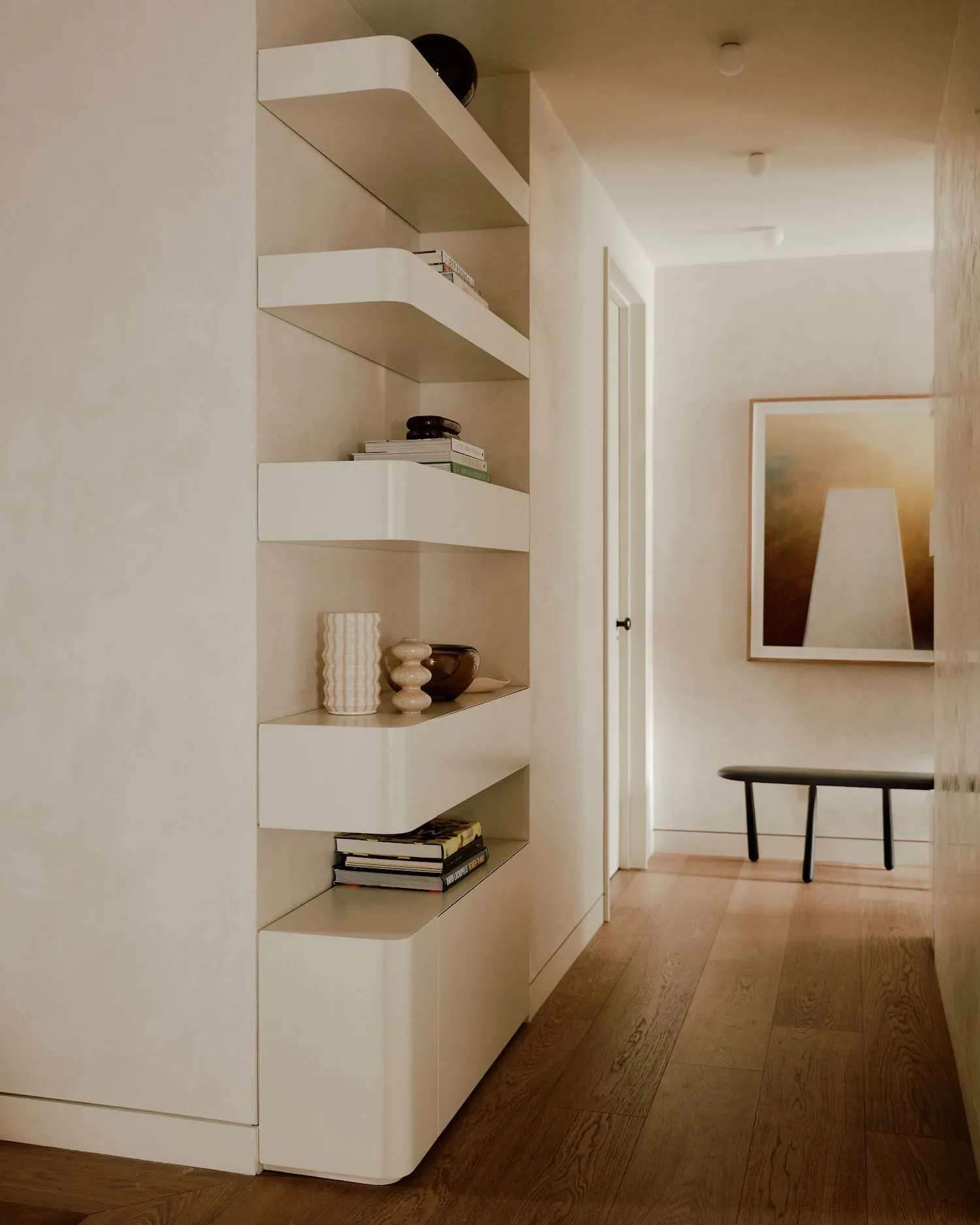 A modern minimalist living room with sleek, white shelves, wooden flooring, and a black bench with a framed abstract artwork.