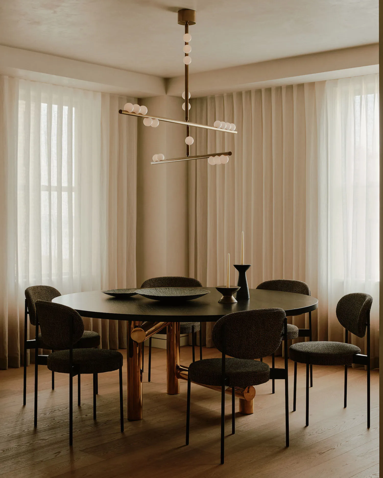 A modern dining room featuring a round wooden table with brass legs and chairs, set against sheer curtains.