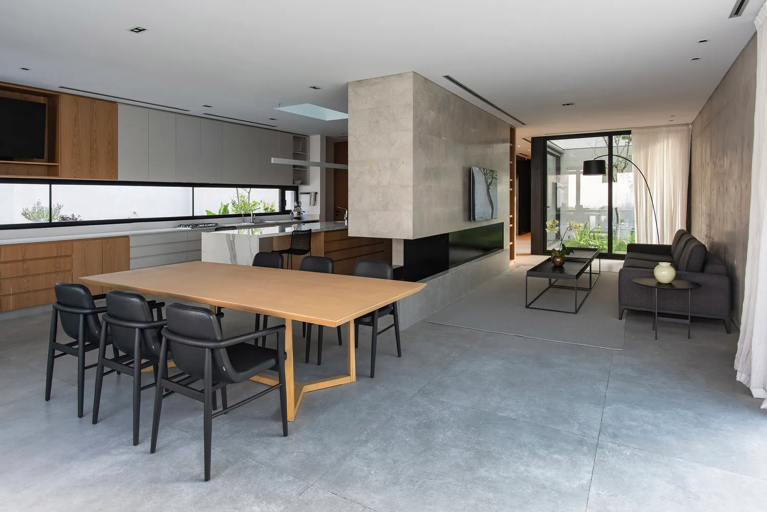 Modern living room with concrete walls and floor, featuring a large table and black chairs. The background includes a kitchen