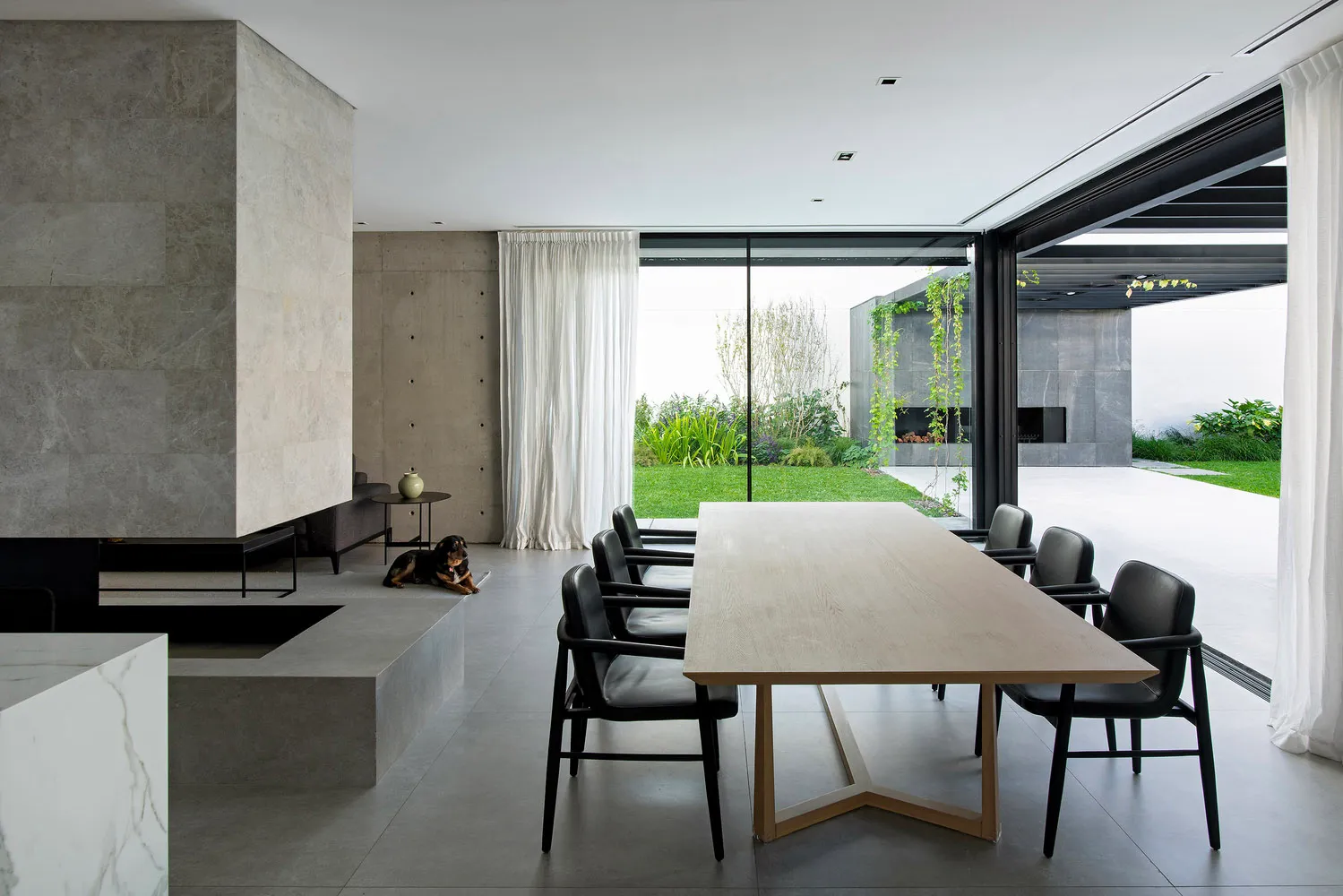 Contemporary residential dining room featuring a long wooden table, black chairs, concrete floors, and a large window overloo