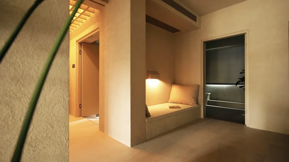 A minimalist interior space featuring a concrete seating area with a cushion and a soft-lit lamp on a beige wall, adjacent to