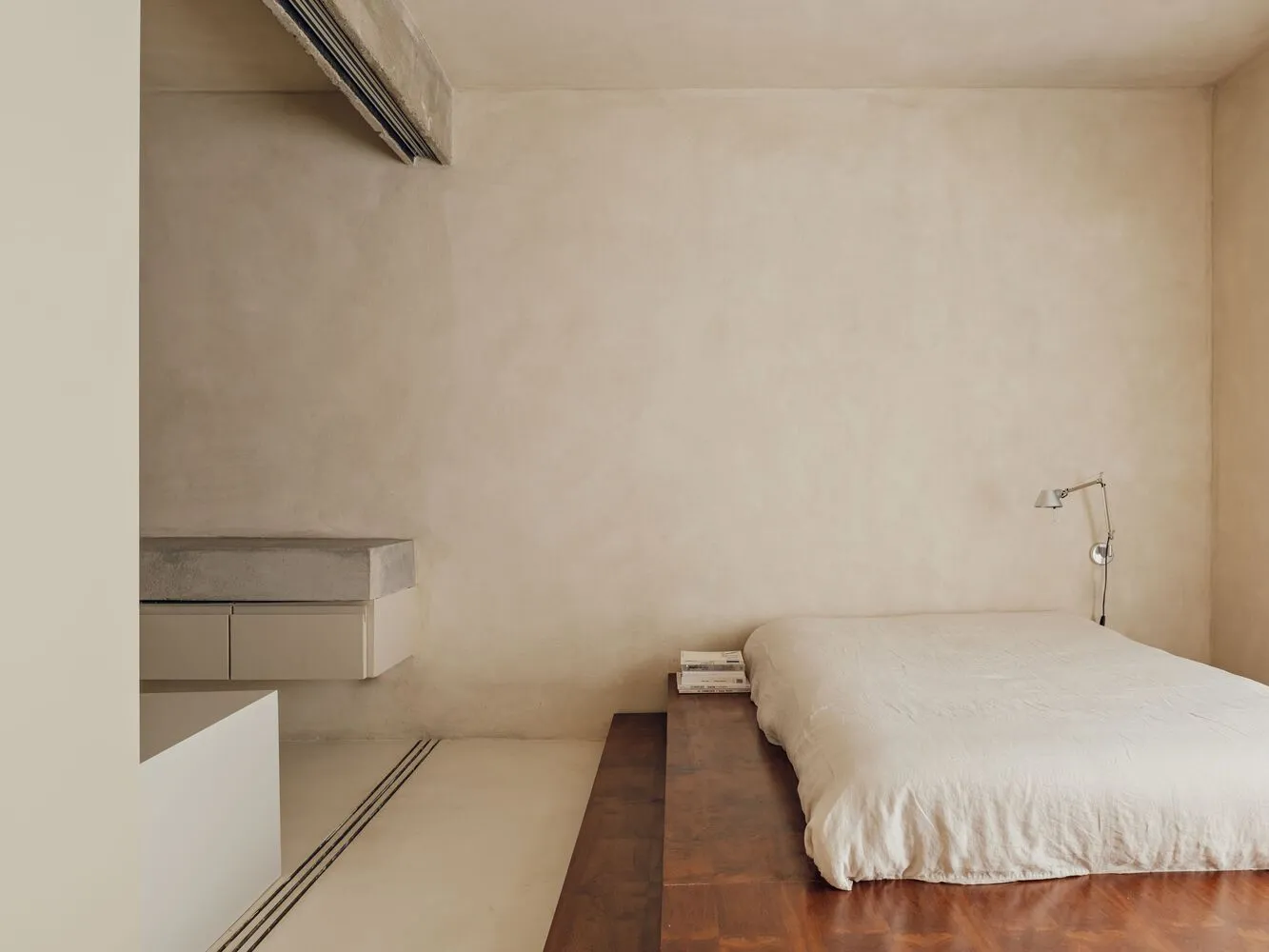 A minimalist bedroom featuring a concrete bed platform on a wooden floor.