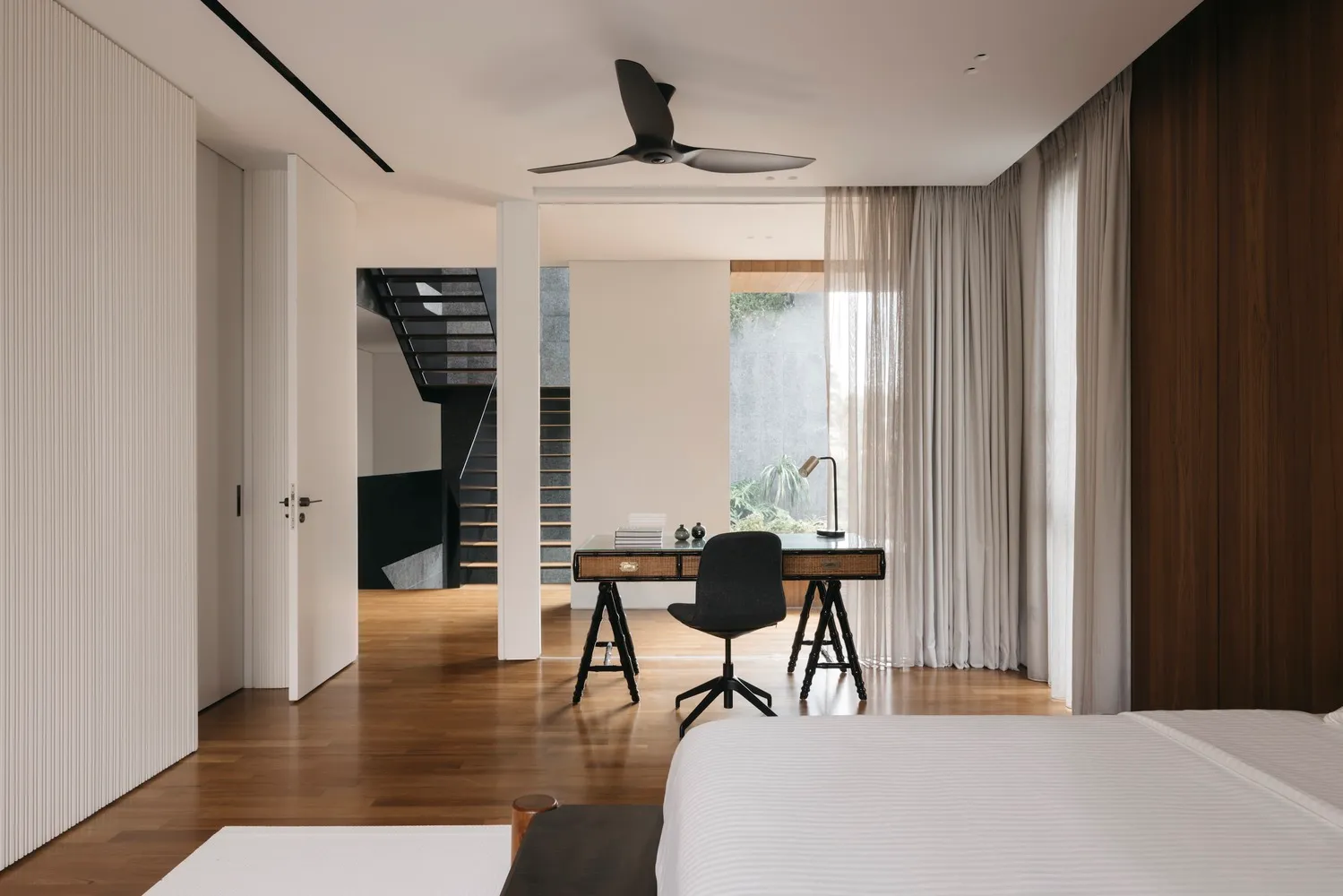 A minimalist residential bedroom featuring wooden flooring, glass windows, fabric curtains, and metal furniture.