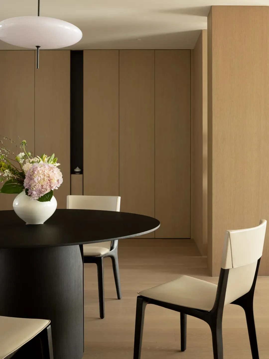 Minimalist dining room with a round wooden table, white chairs, and a white vase with flowers.