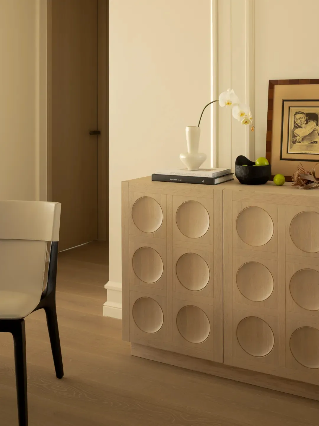 A modern interior design featuring a wooden cabinet with circular cutouts, an orchid, green apples, and a framed artwork.