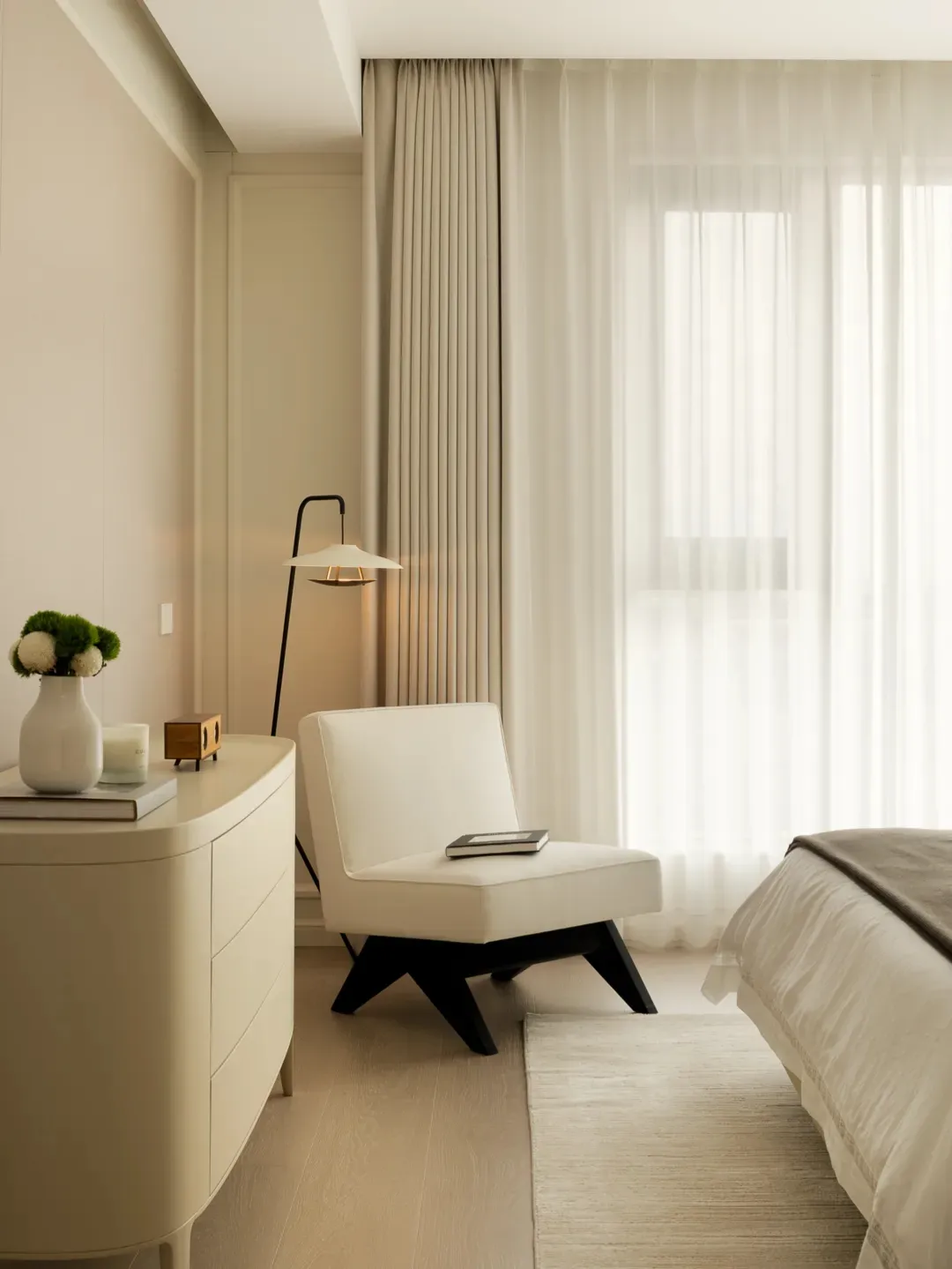 A modern minimalist bedroom featuring a minimalistic white armchair with a black frame sitting by a window with sheer curtain