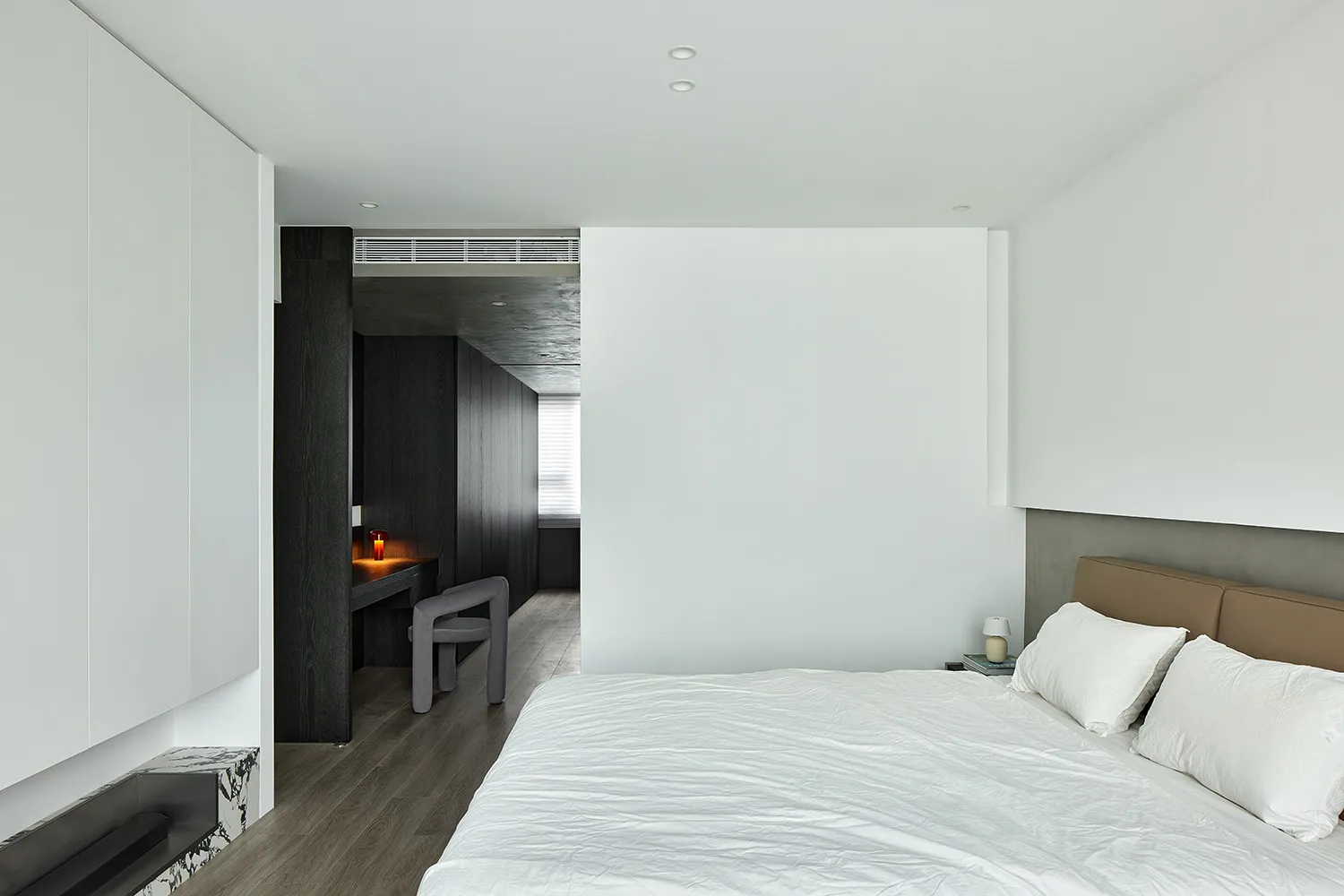 A minimalist bedroom featuring a white clothed bed, with white walls and a mix of dark and light wooden textures.