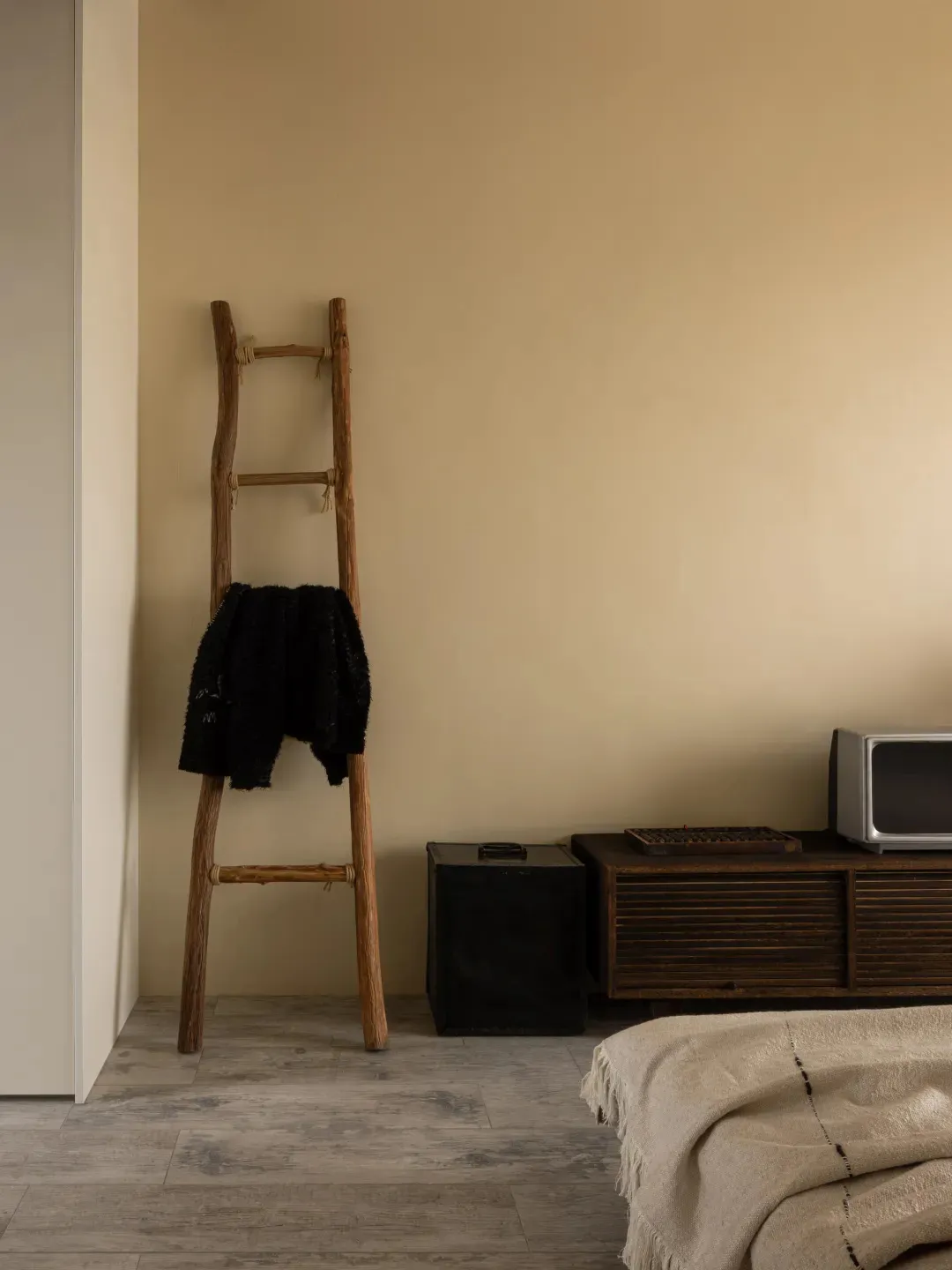 A minimalist bedroom featuring a wooden ladder with a blanket on it, against a beige wall, with rustic wooden furniture and a
