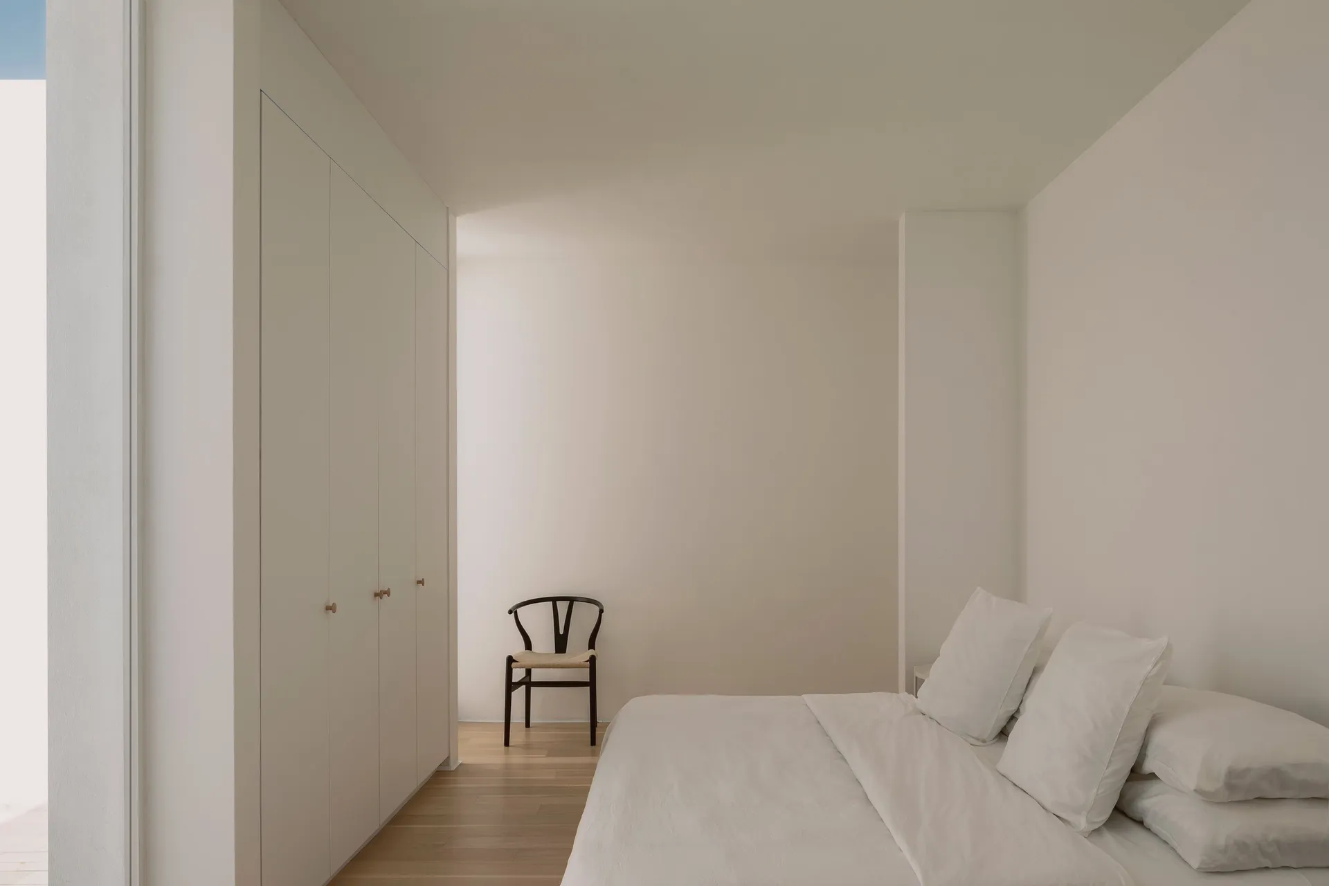 A minimalist bedroom with white walls, a wooden bed, and a simple chair.