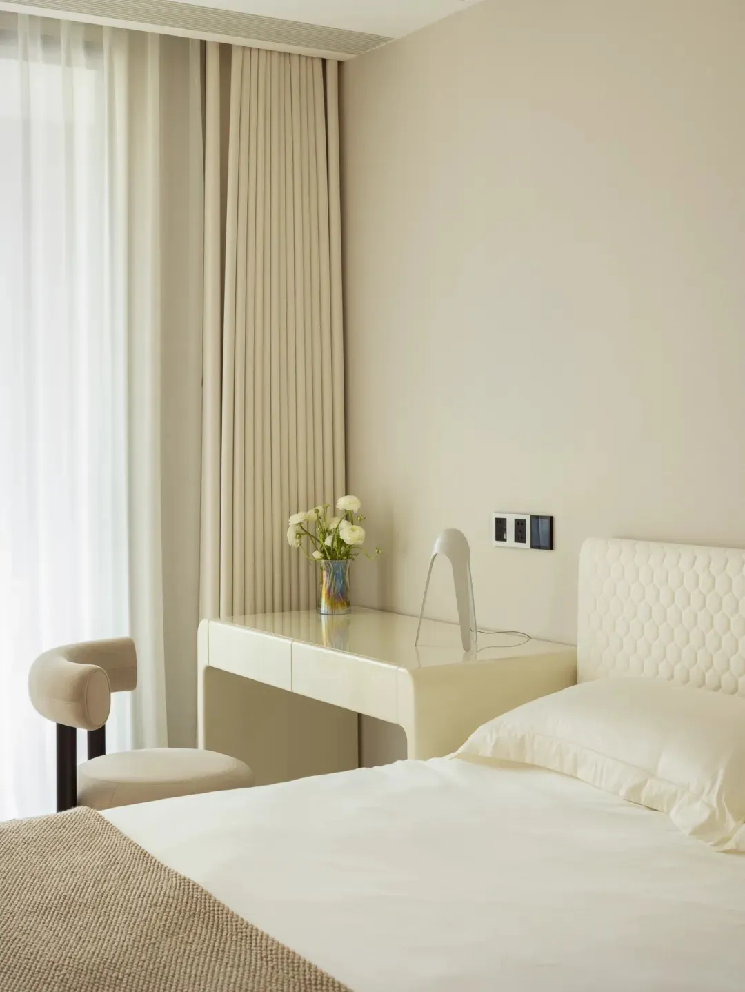 A minimalist bedroom featuring a white fabric bed against a light beige wall, illuminated by natural light filtering through
