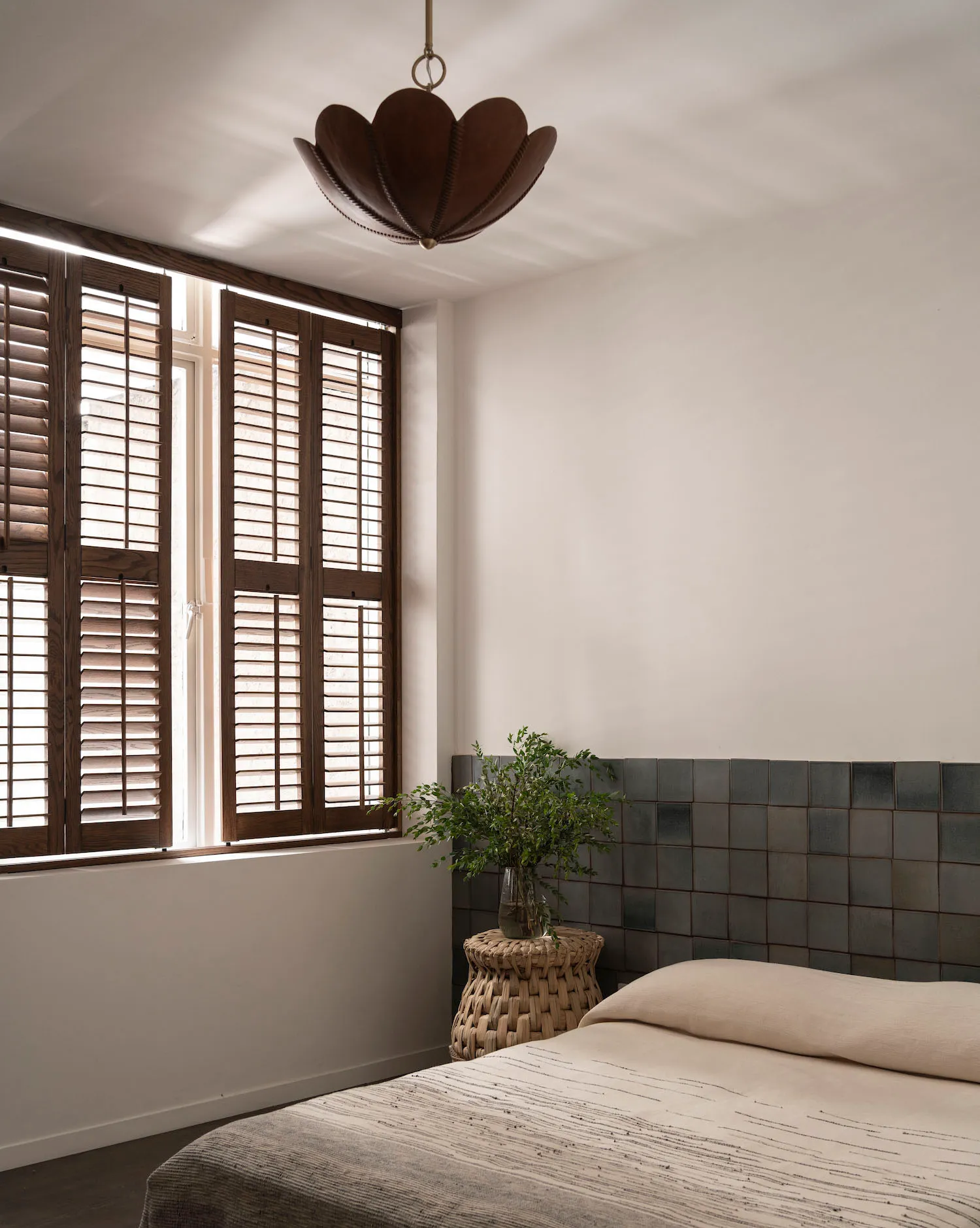 A minimalist bedroom featuring wooden fixtures, concrete tiles on the wall, and a small potted plant on a woven stool.