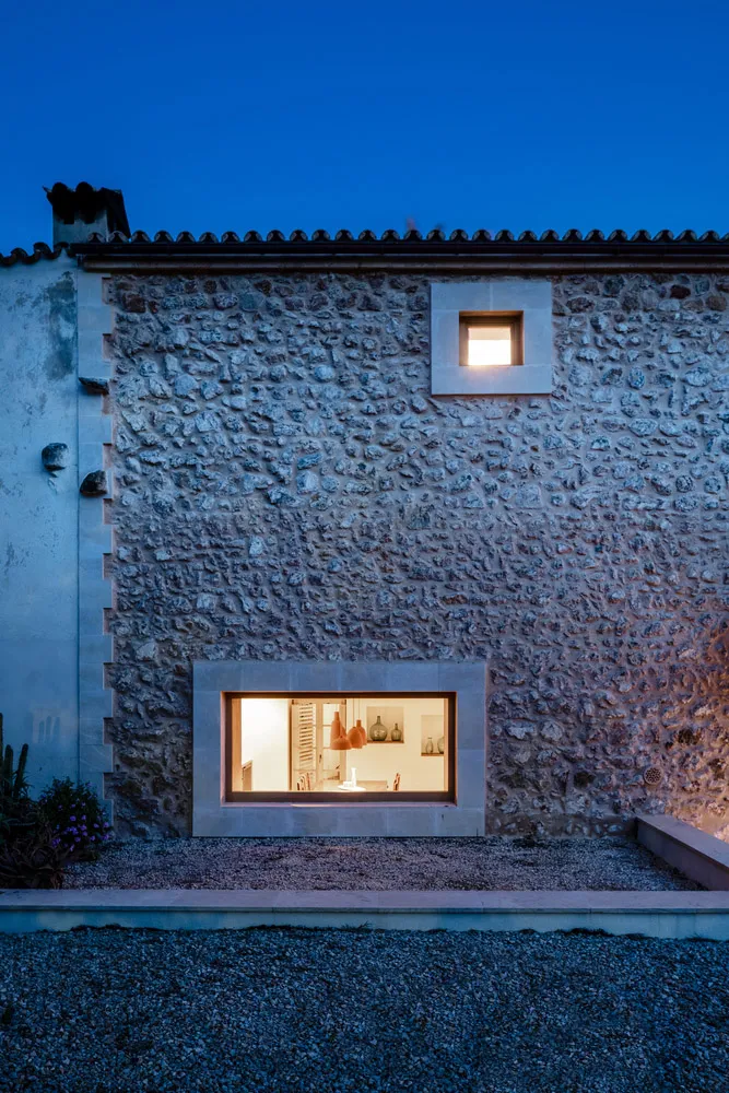 A stone house exterior illuminated at dusk, showcasing a small window and a large stone-framed window with a view into the in