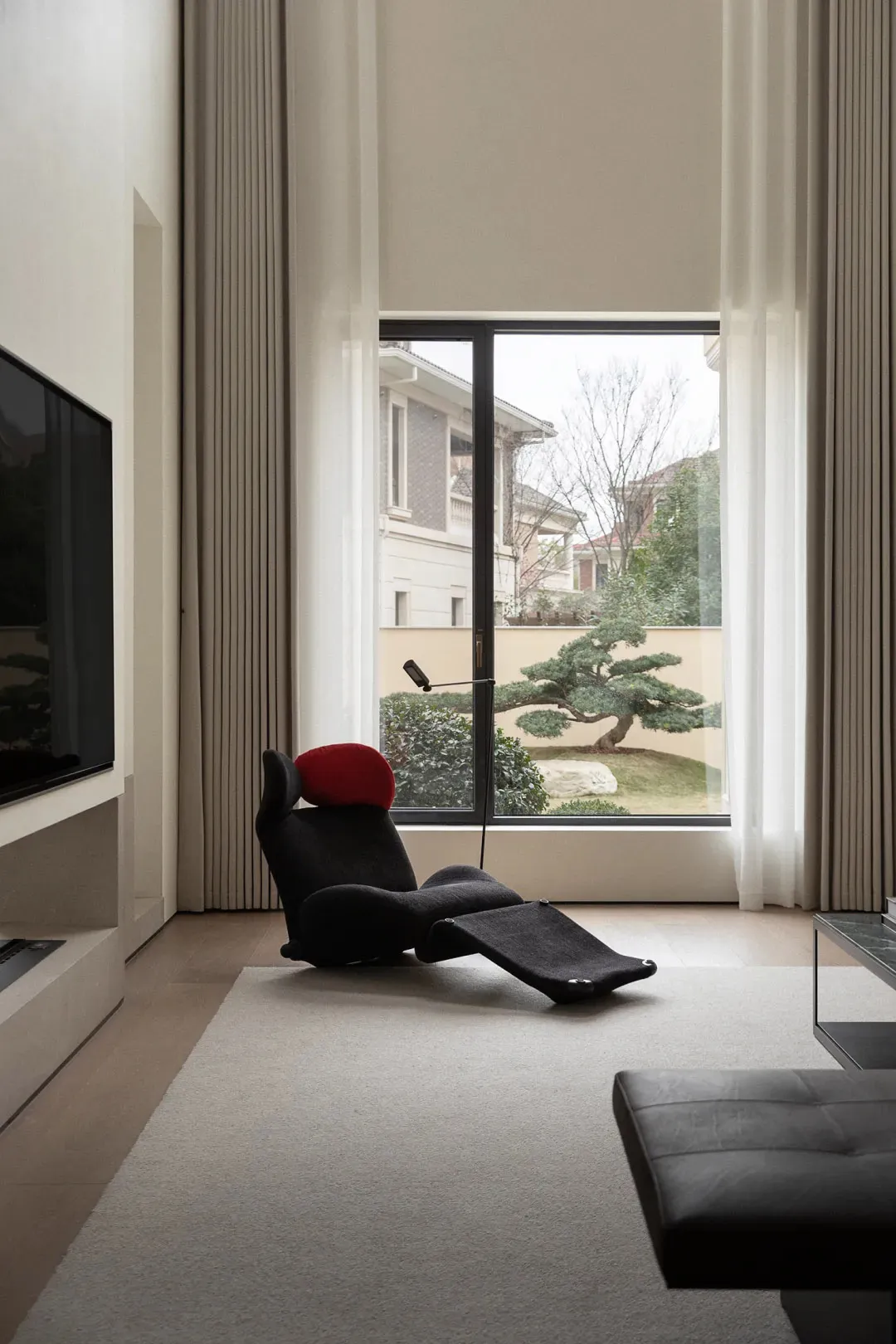A minimalist living room with a textured cloth recliner chair, a flat-screen television, and a large window with white curtai