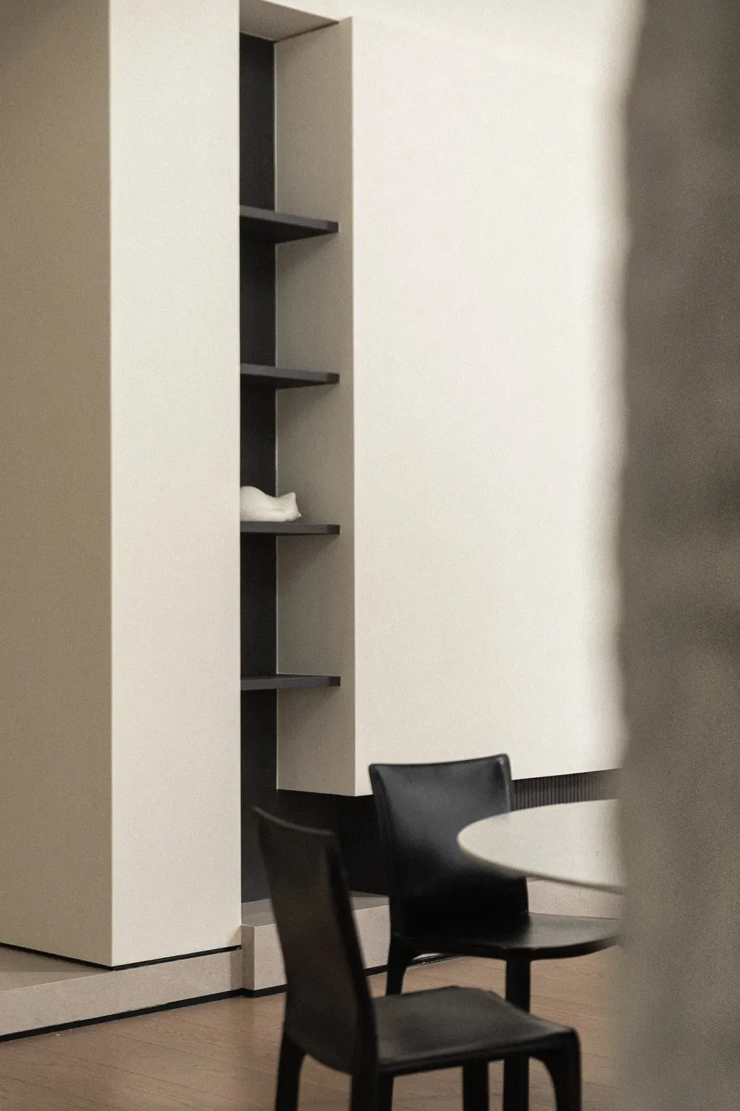 Two black chairs with a round table in front of a minimalist shelving unit with wood shelves and a white wall background.